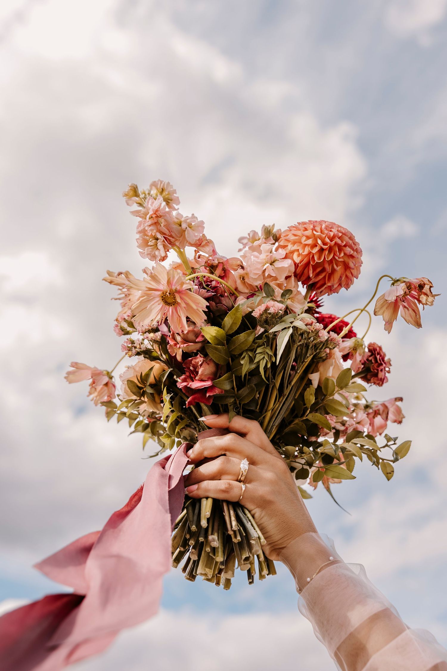 Hands holding up a pink and coral wedding bouquet against a cloudy sky.