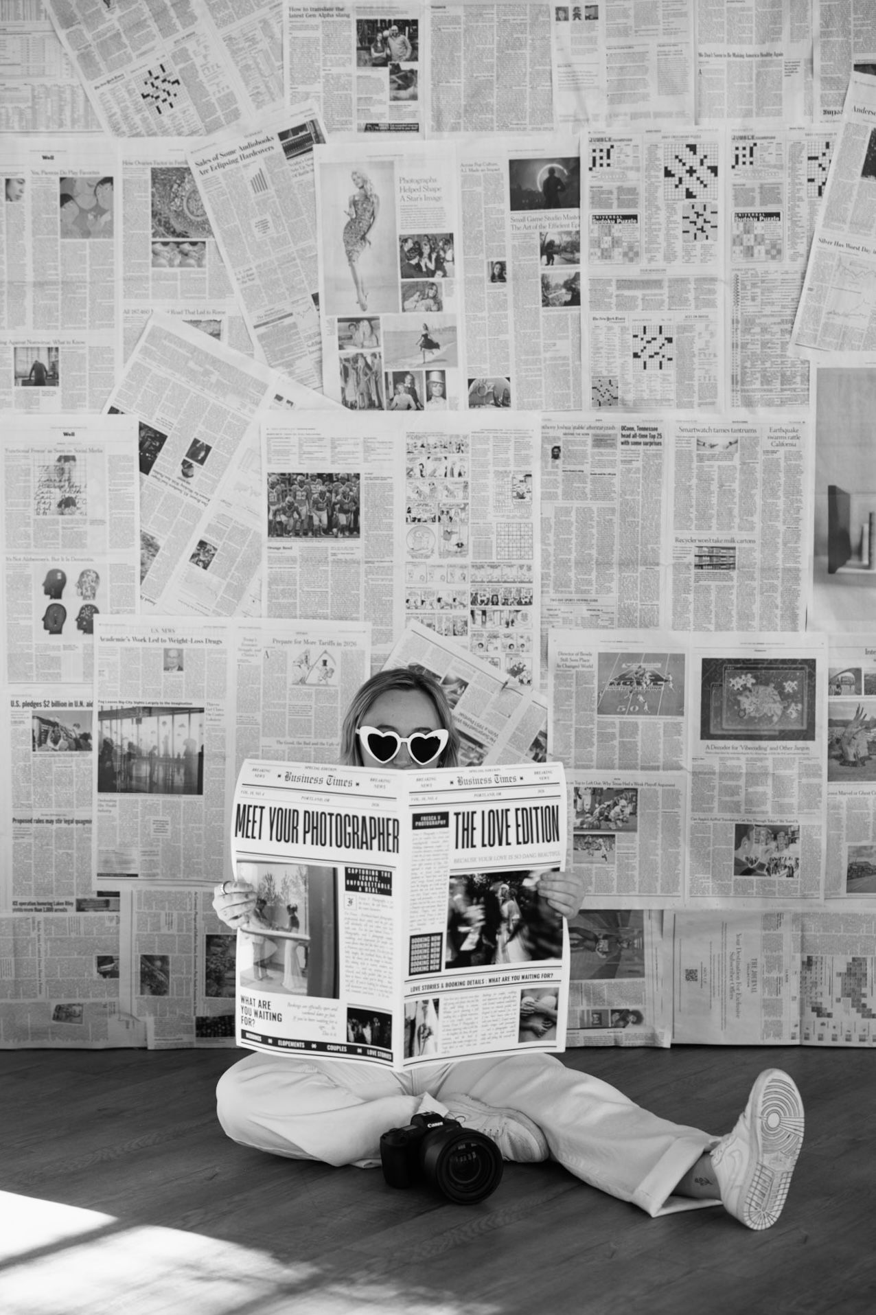 Black and white photo of a person sitting on floor holding a newspaper in front of a wall covered with newspaper pages.
