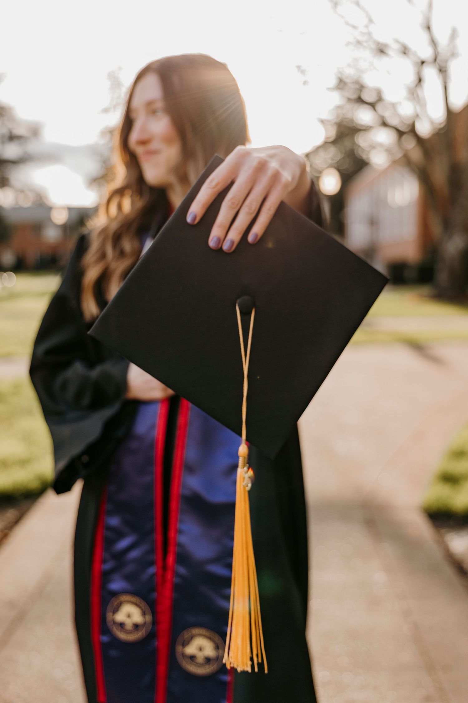 Graduation cap with tassel held up against sunlit campus background.
