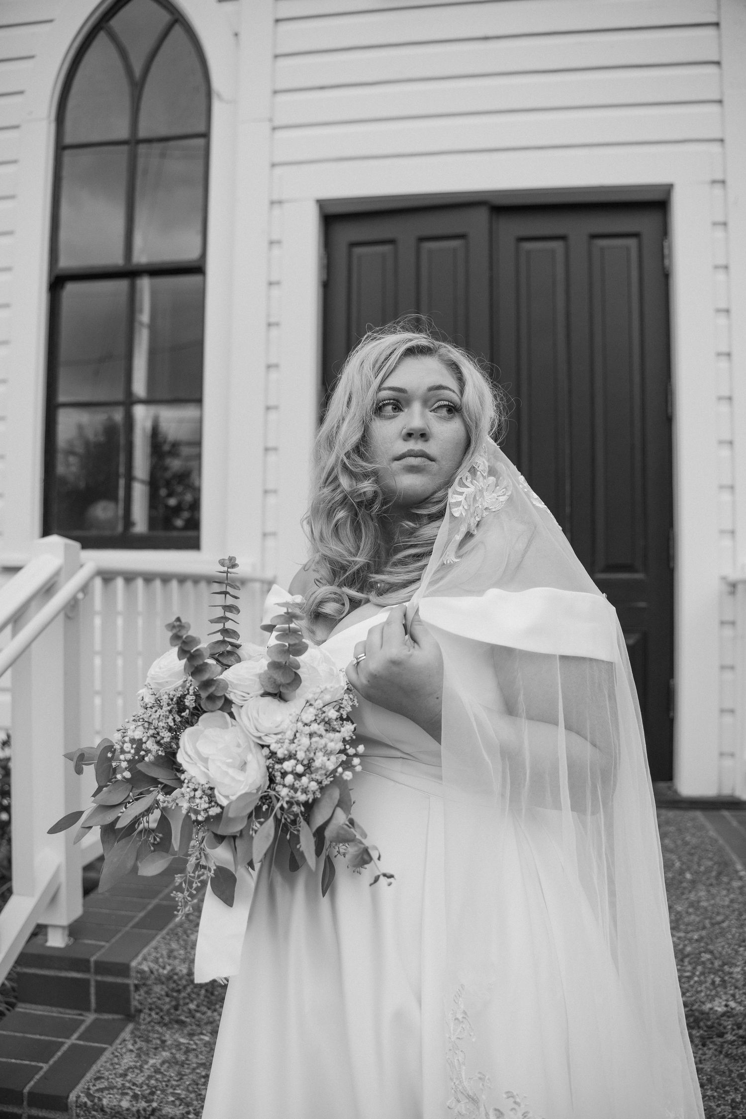 A bride in a flowing veil poses with her bouquet on the steps of a white church.
