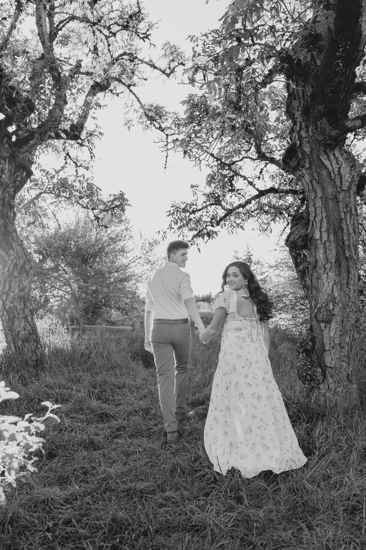 A romantic couple holds hands under a large tree in a black and white photograph.