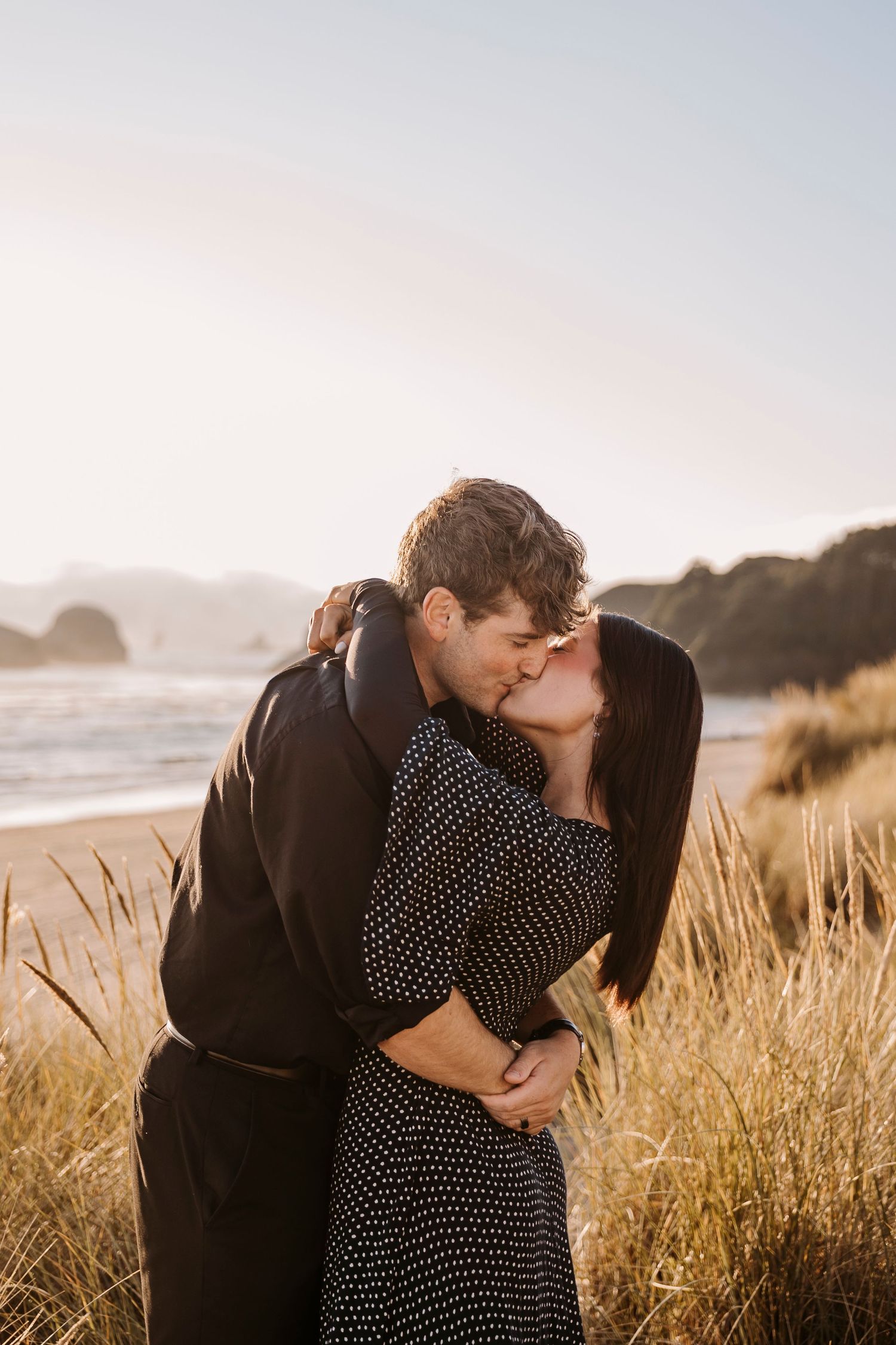 A couple shares a kiss on a beach at sunset with coastal rocks in the background.