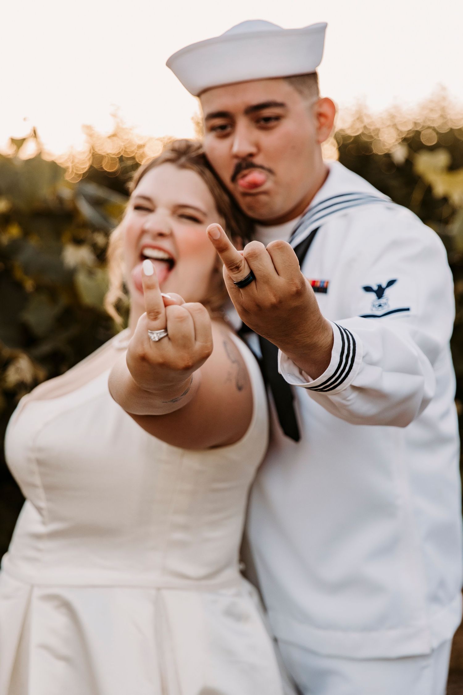 A sailor and bride in white make playful gestures at the camera during their outdoor wedding photoshoot at sunset.