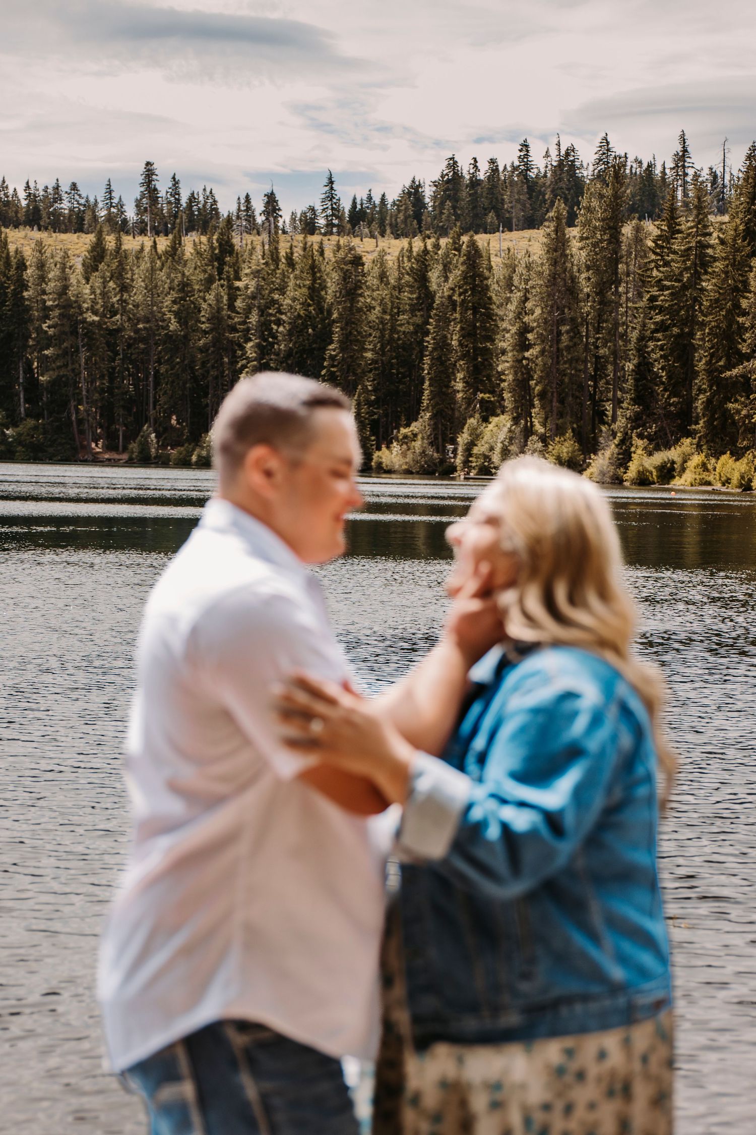 A couple embraces by a serene mountain lake surrounded by pine forests.