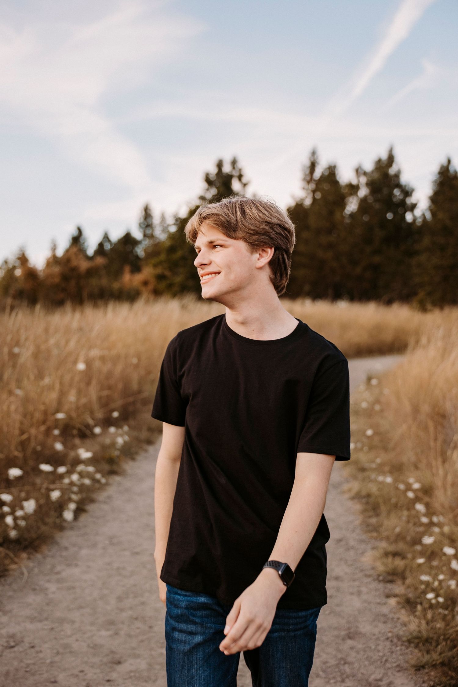 Casual outdoor portrait on dirt path with evergreen trees and golden grass in background.