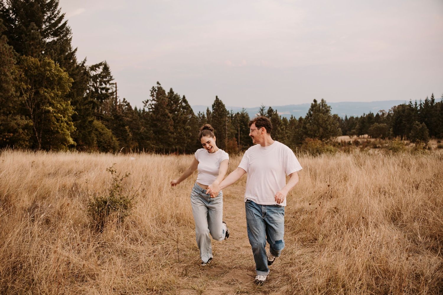 A couple walks through a golden field at sunset with tall evergreen trees in the background.