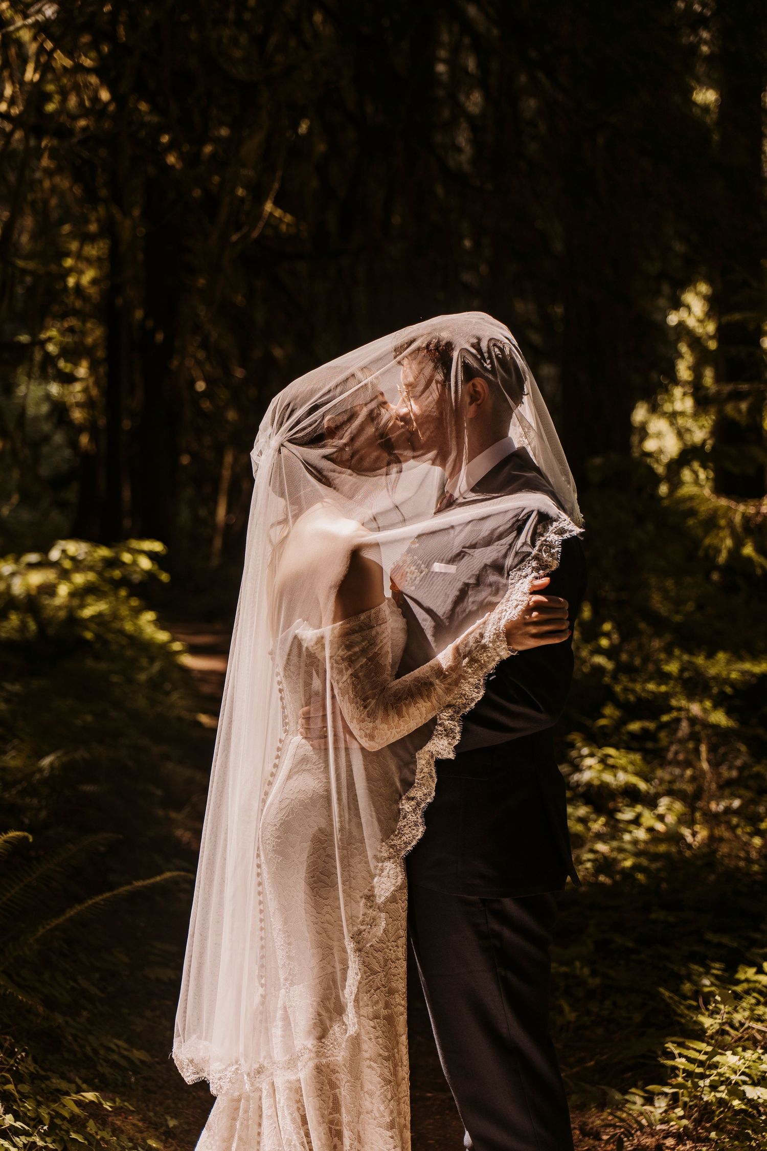 Bride and groom share a romantic kiss under a sheer lace veil in a sunlit forest setting.