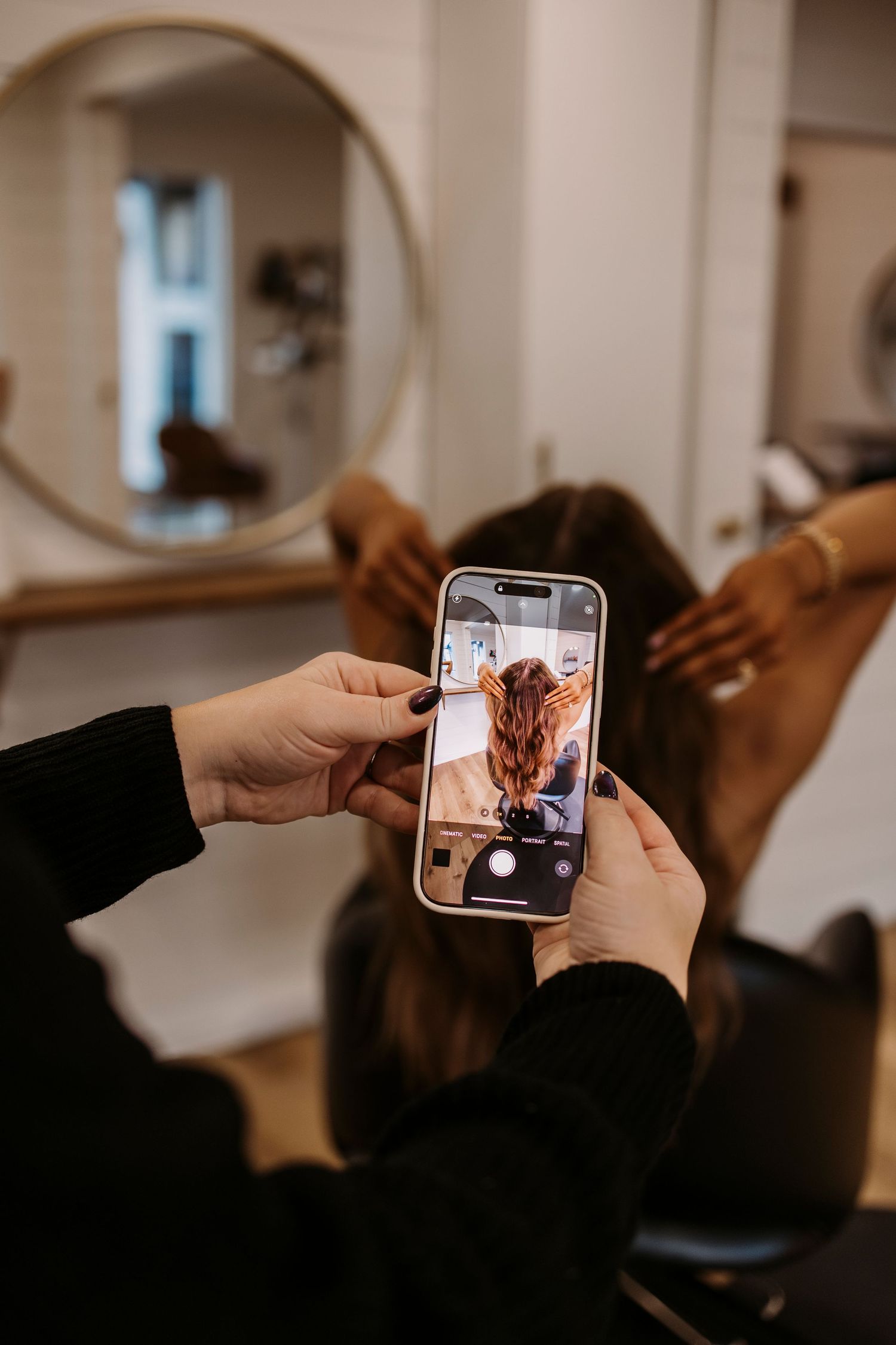 Taking photos of hairstyling process with smartphone in salon mirror reflection.