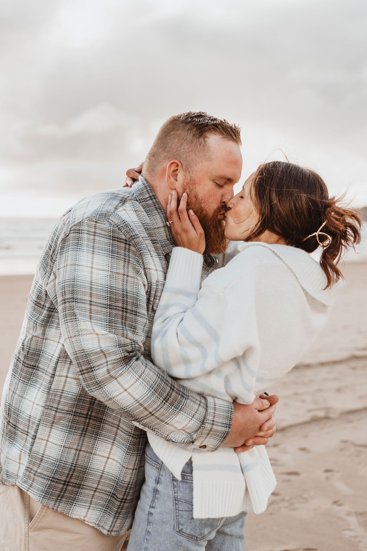 Intimate beach engagement session with couple in casual attire on sandy shore.