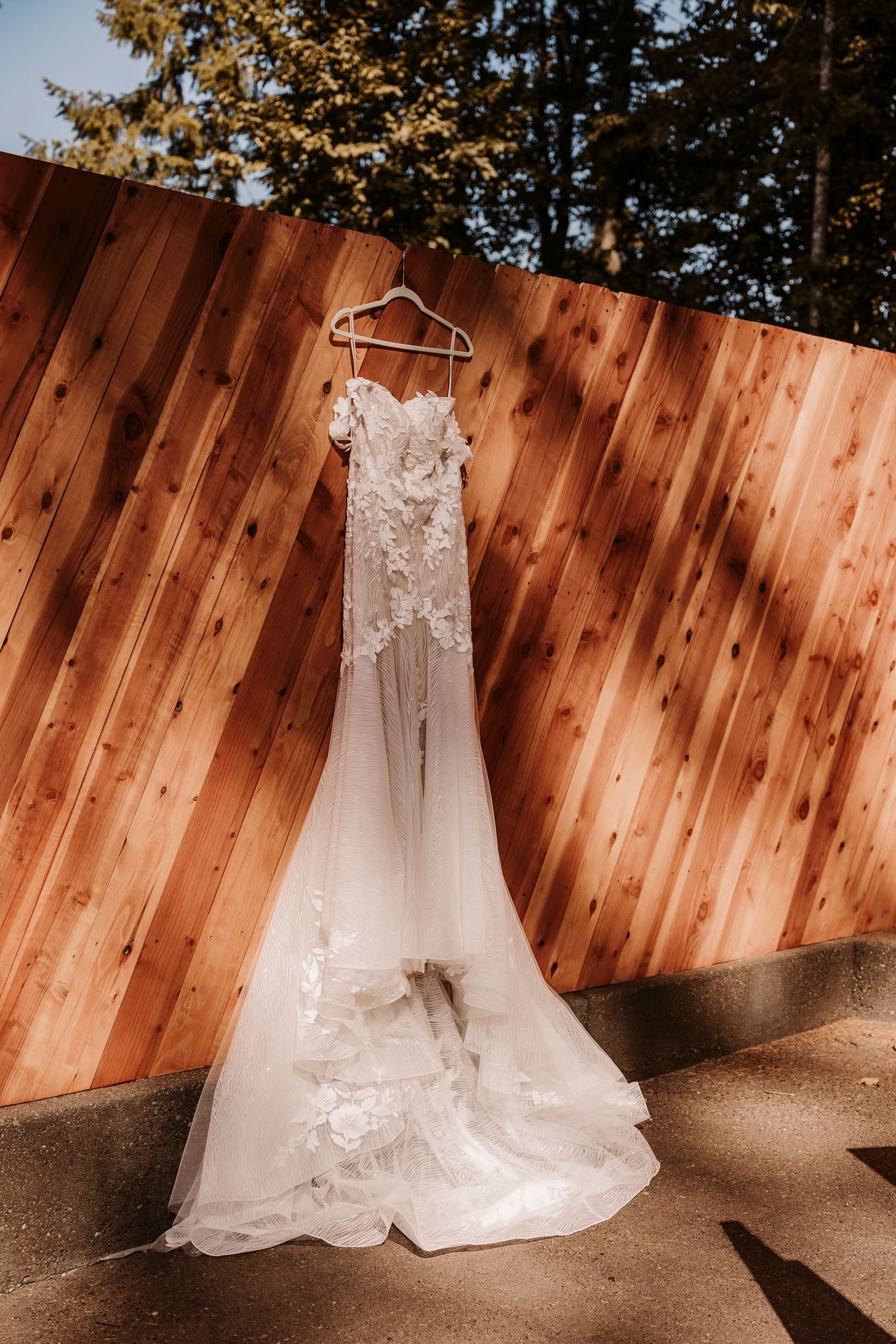 White lace wedding dress with floral appliques hanging against a wooden fence in sunlight.
