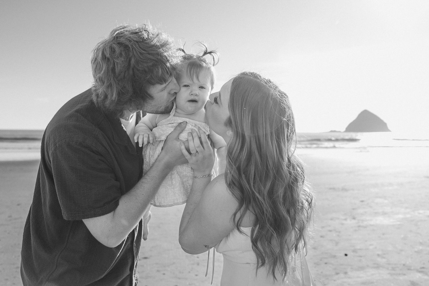 A family shares a tender moment together on a beach with Haystack Rock in the background.