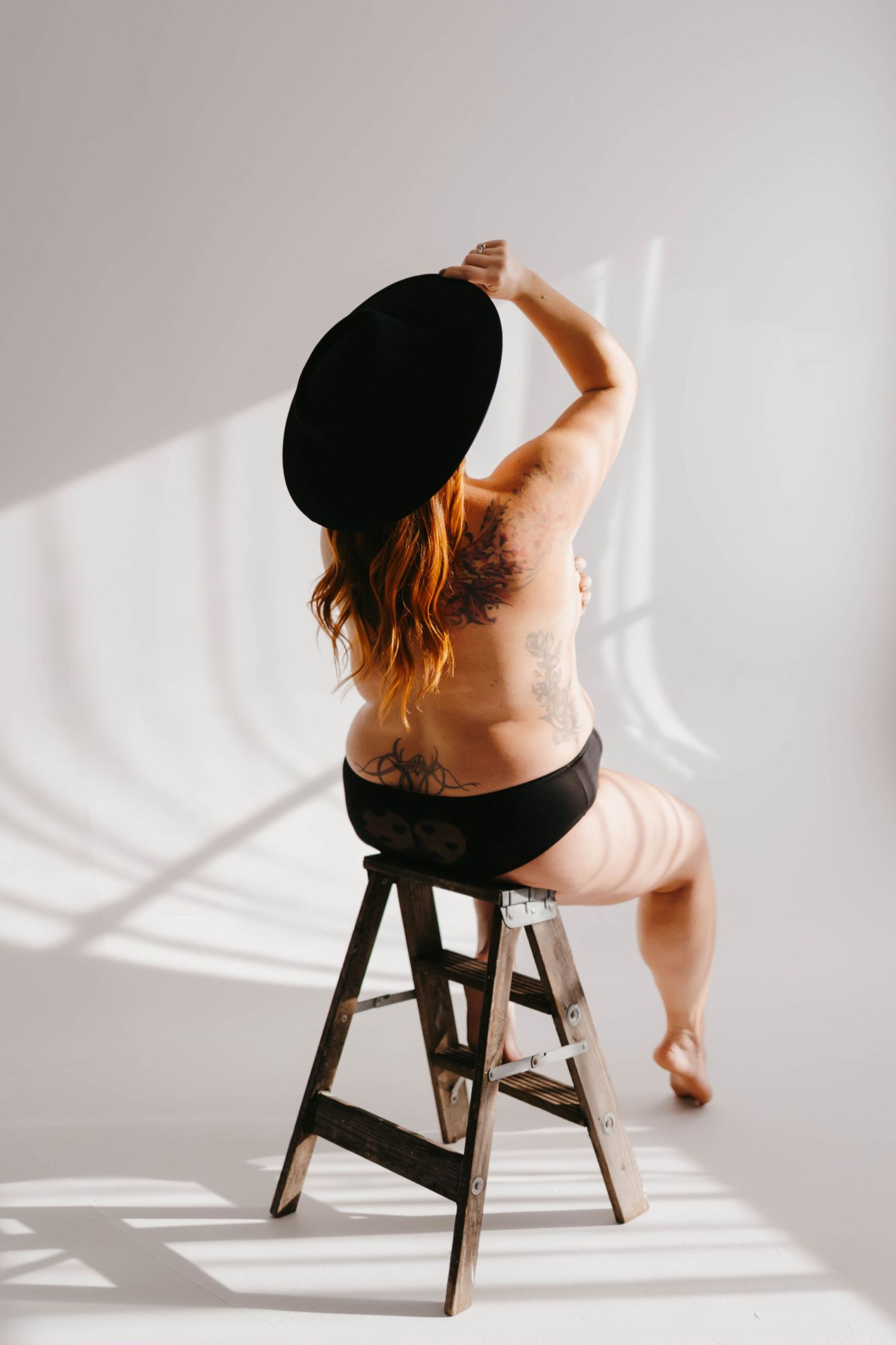 Artistic boudoir photo series showing figure posing on wooden stool in natural window light.