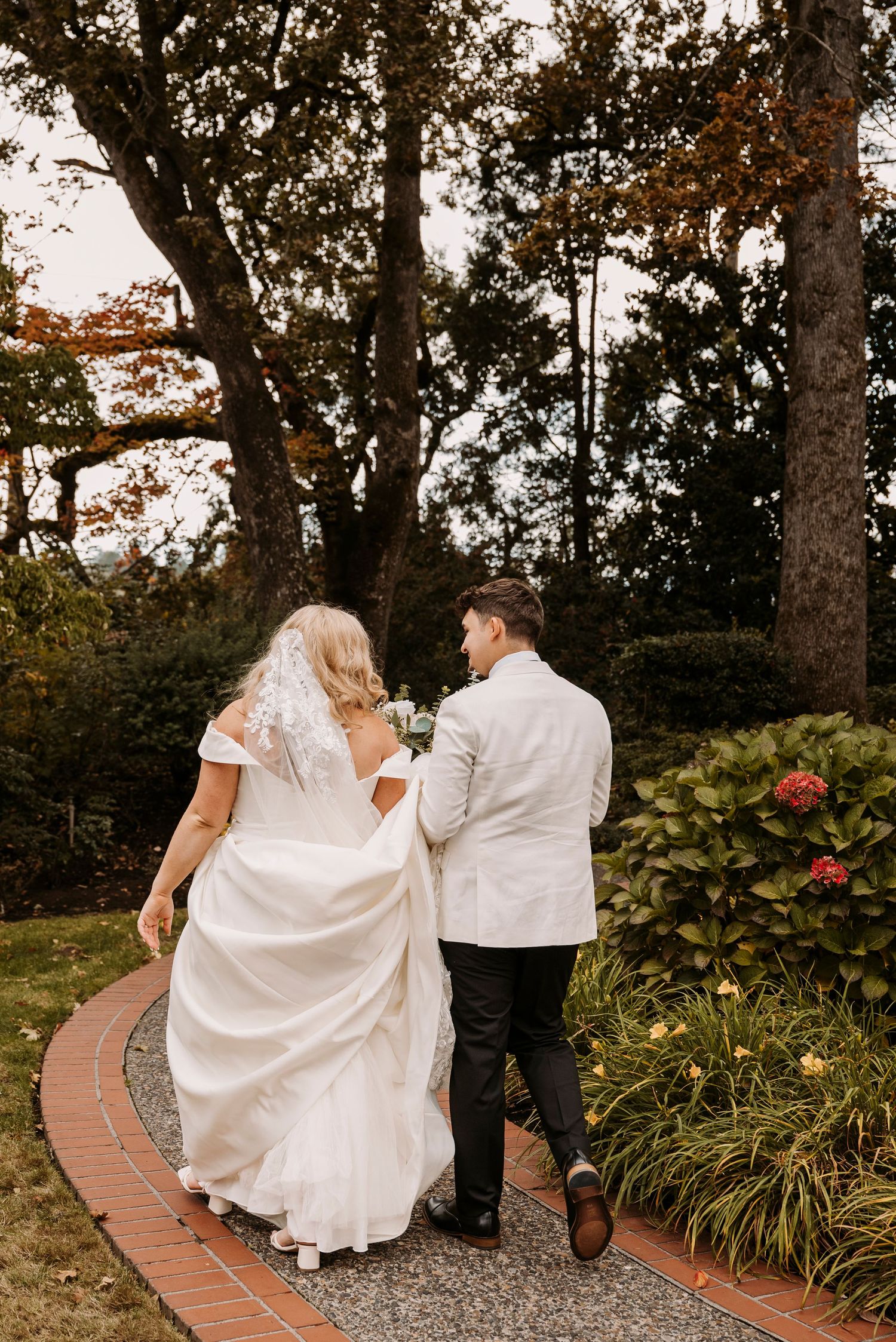 Newlyweds walk along curved brick path outside white chapel.