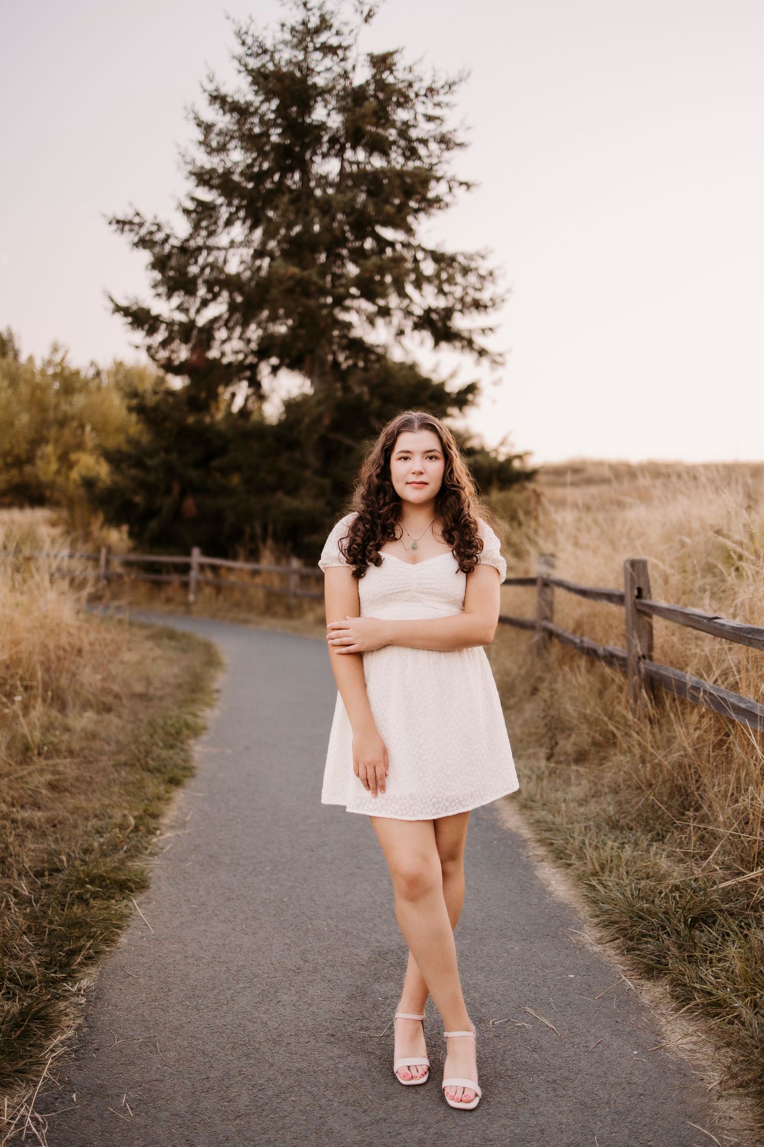 Someone posing on a paved path with evergreen trees and a rustic fence in warm evening light.