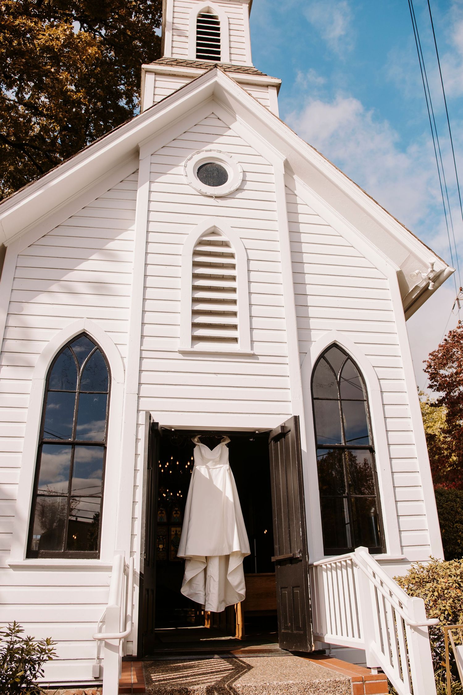A classic white chapel with Gothic windows displays a wedding dress hanging in its doorway.