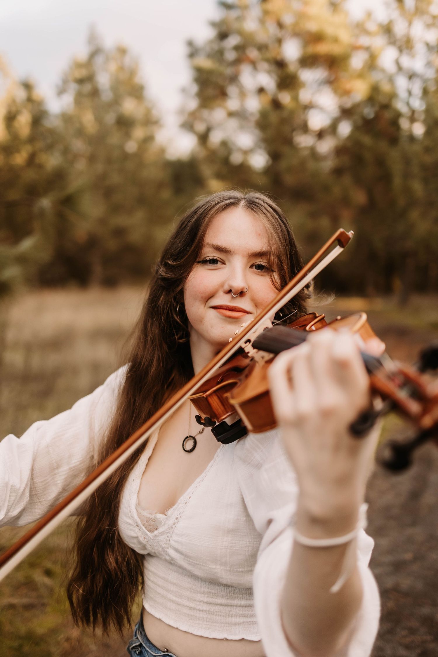 A musician plays violin outdoors in a white top and jeans during golden hour.