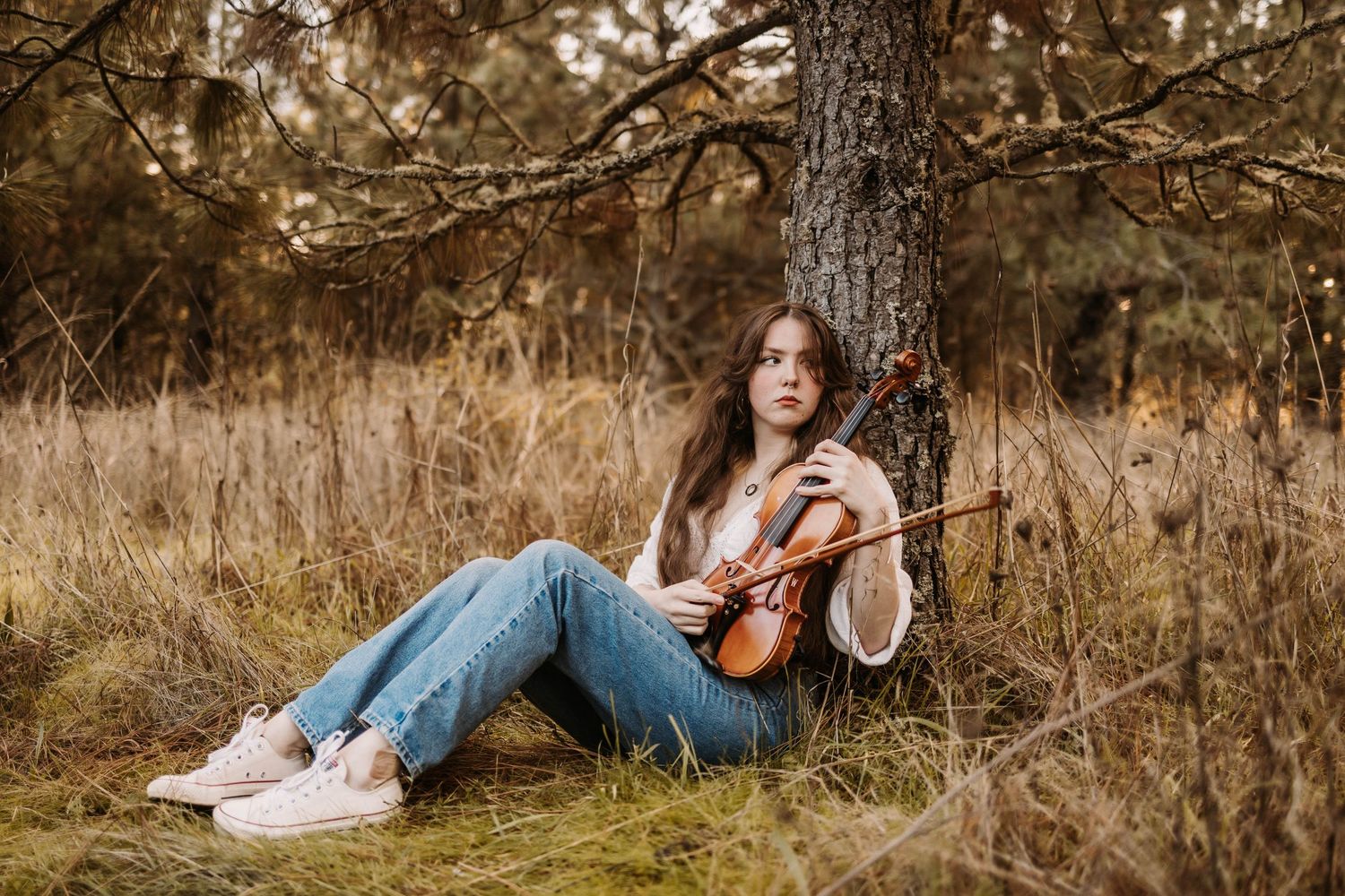 A violinist sits casually against a tree trunk in a natural setting wearing casual denim.