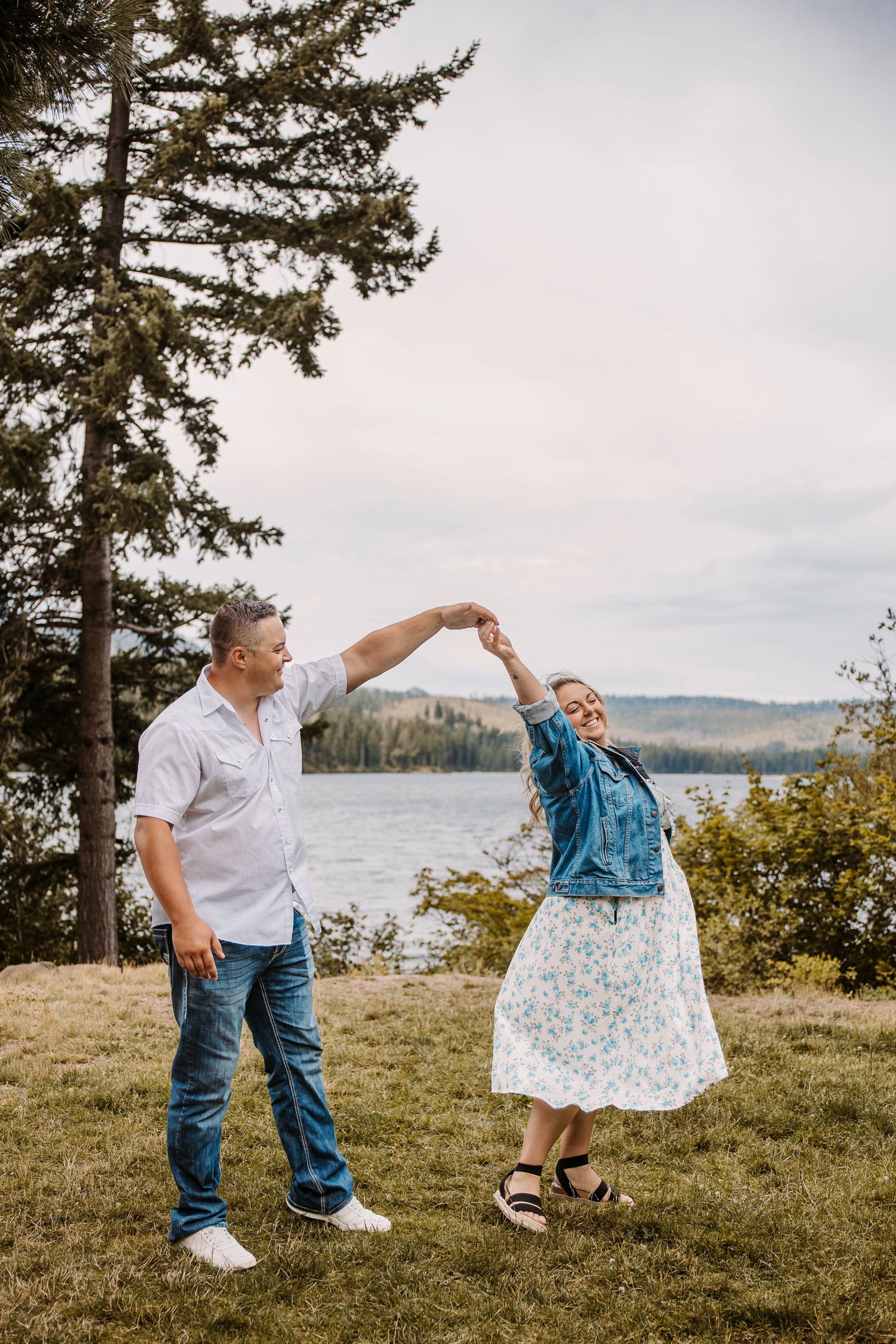 A couple dances playfully by a lake with evergreen trees in the background.