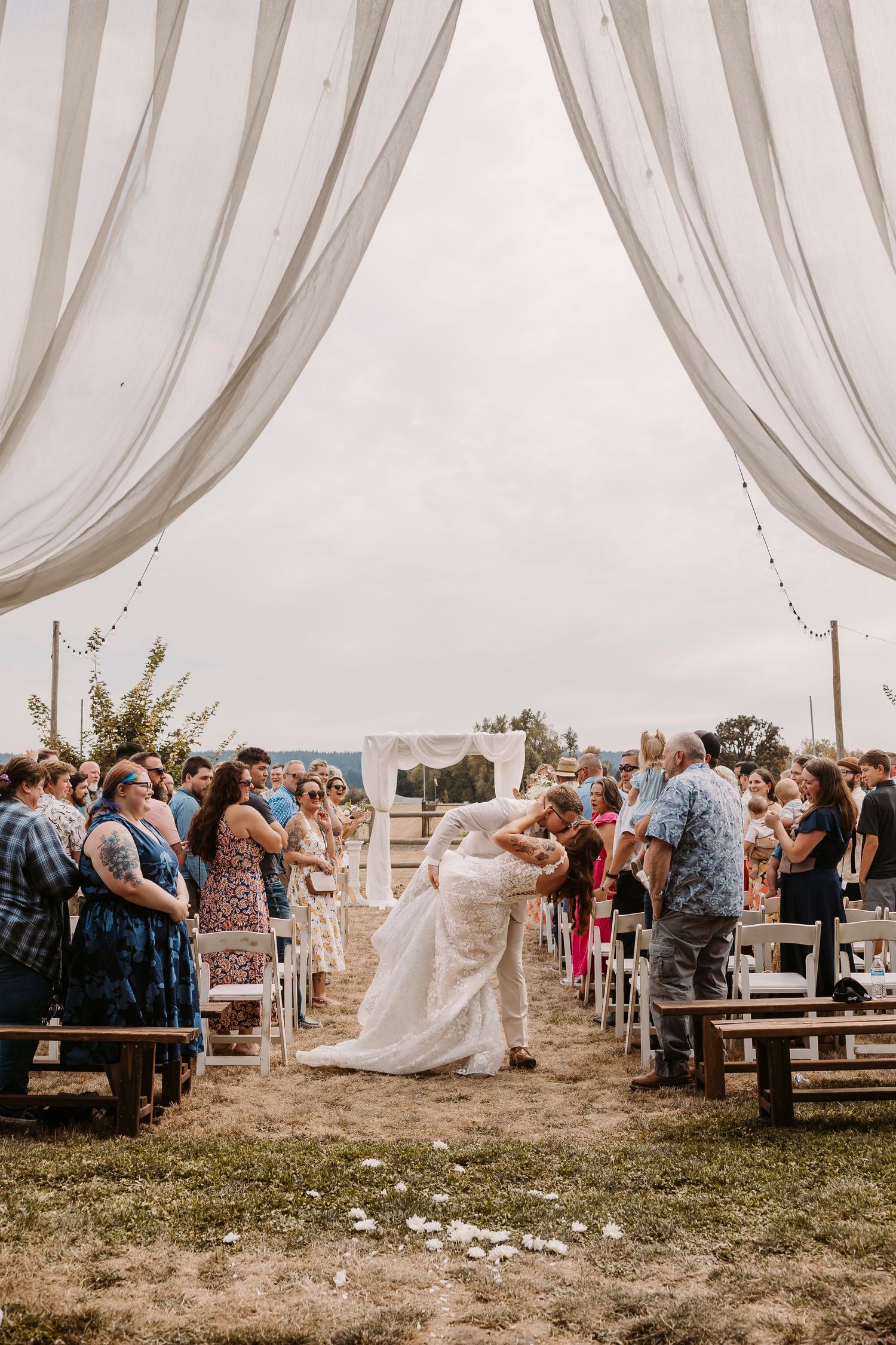 Wedding guests gather outdoors under elegant white draped fabric decorations during a rustic countryside ceremony.