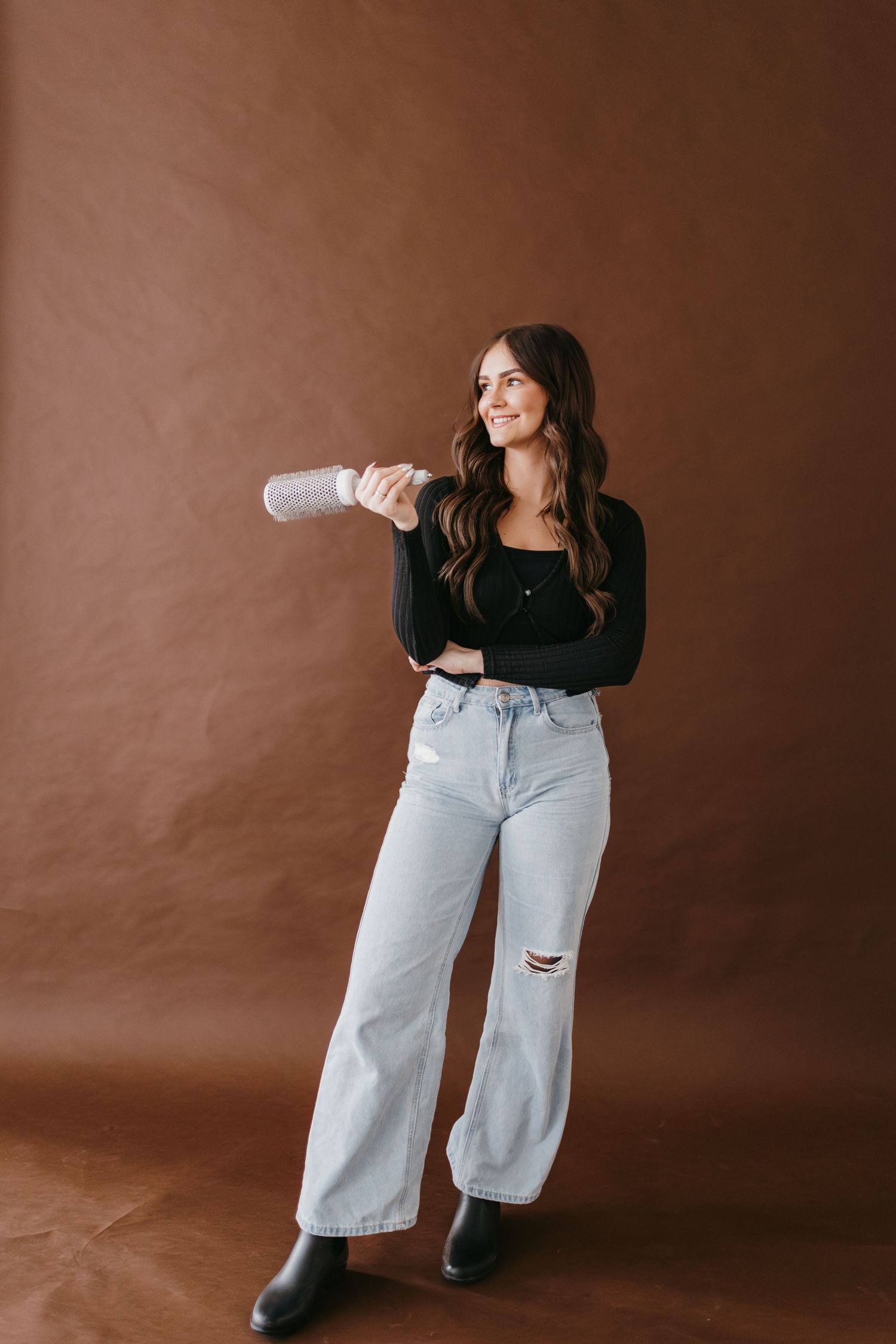Model in black top and light wash wide-leg jeans poses against brown backdrop in fashion photoshoot.