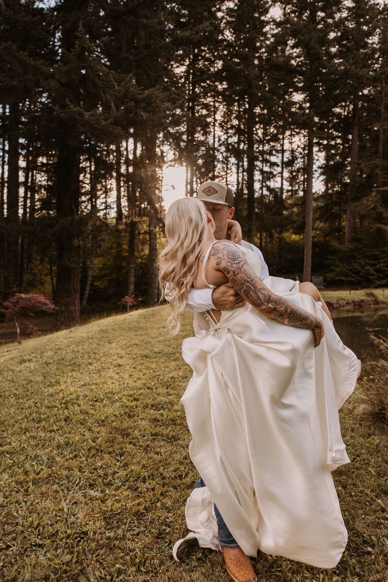 Groom carrying bride in white gown through sunlit forest clearing during golden hour wedding photos.