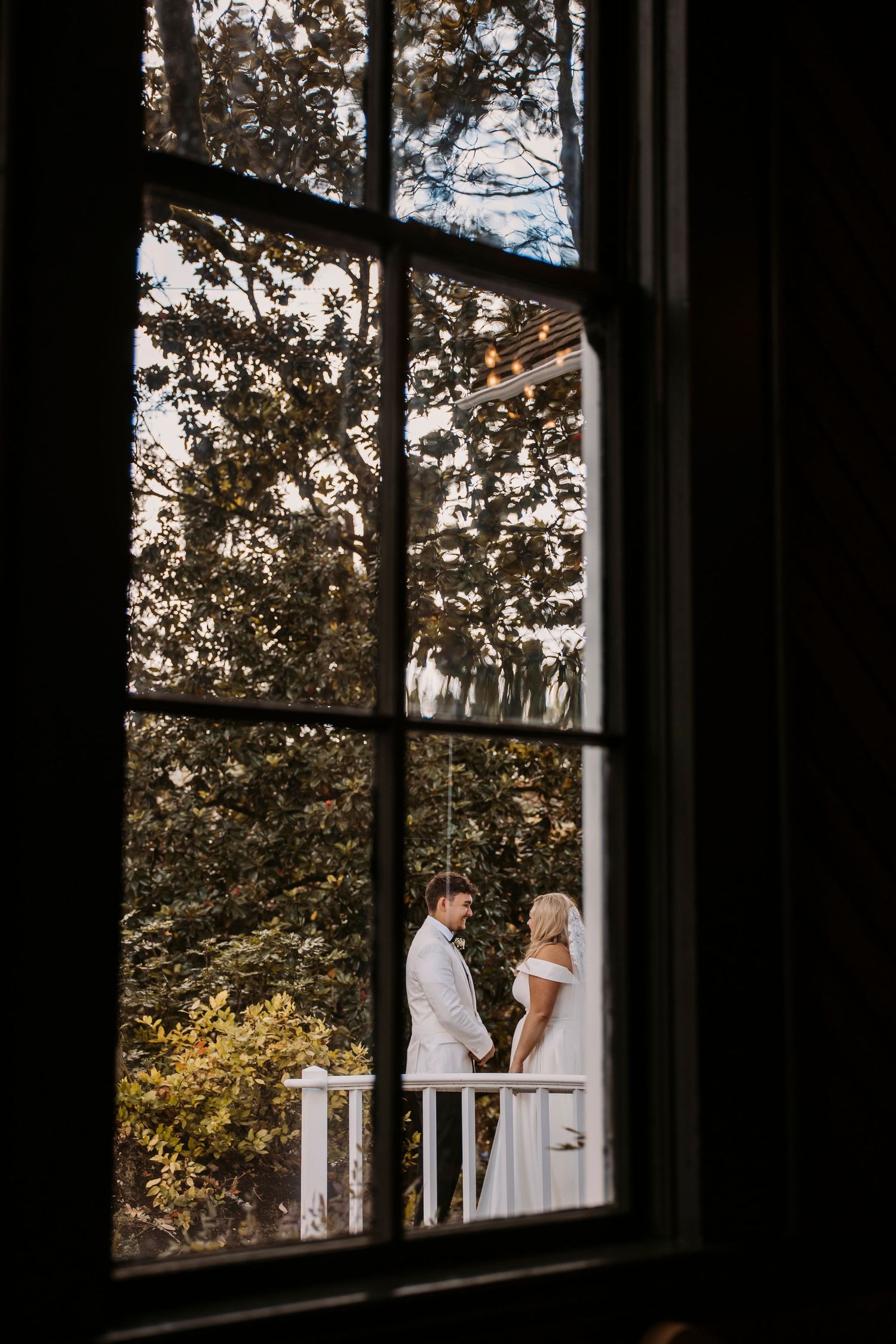 A romantic wedding moment captured through a window frame with trees in the background.