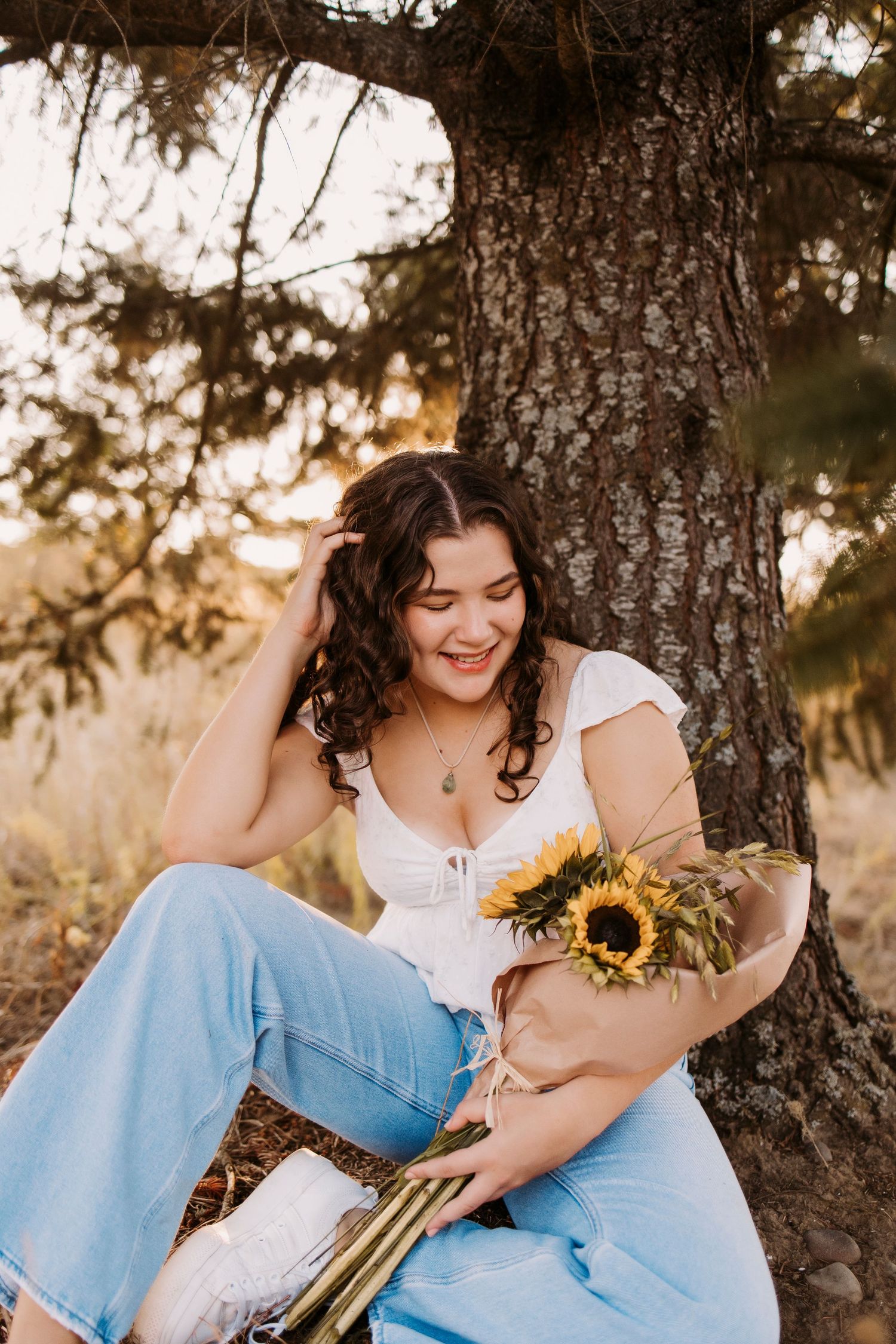 Someone sitting under a tree at sunset holding bright yellow sunflowers while wearing casual denim.