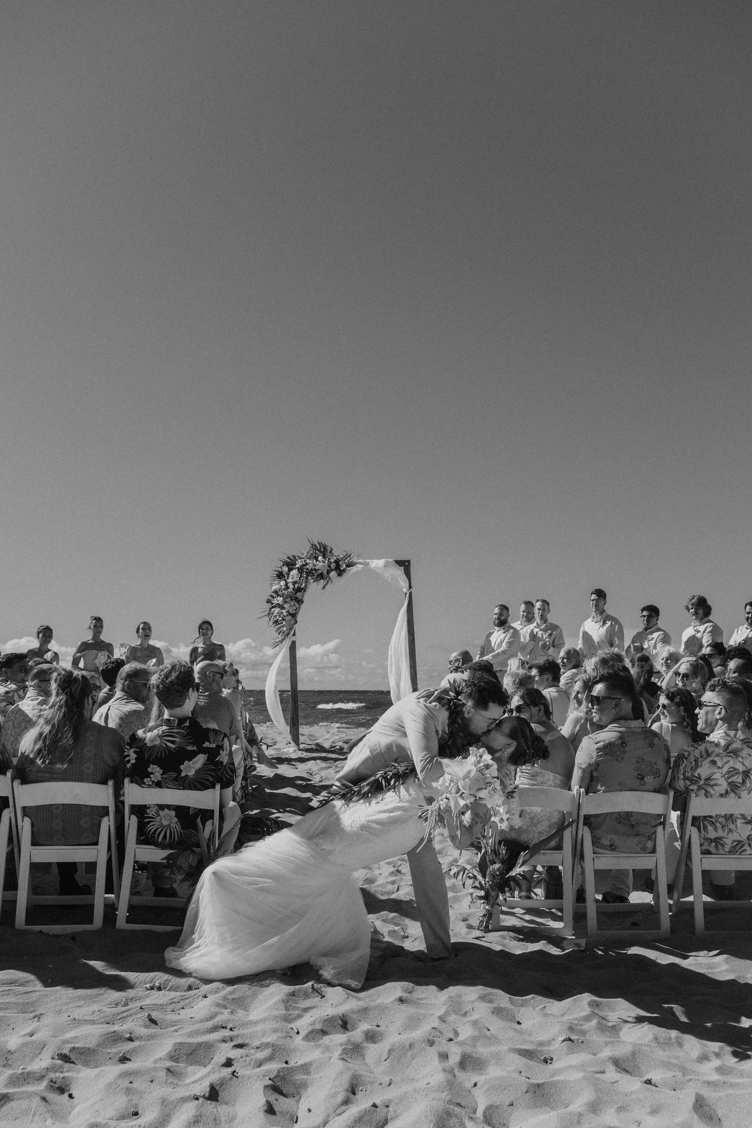 Black and white beach wedding ceremony with bride and groom kissing at the altar, guests seated on white chairs by the ocean.