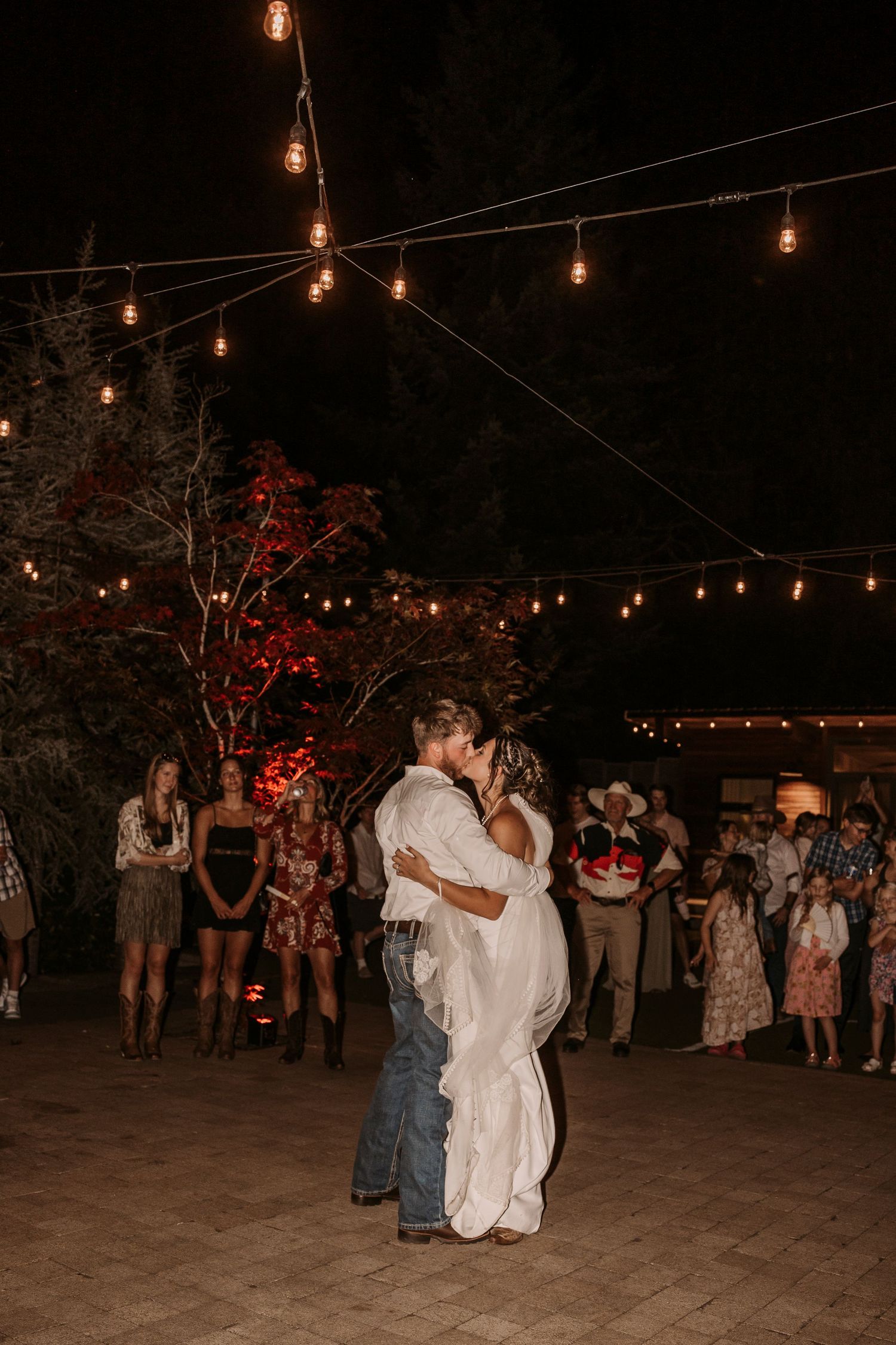 Wedding reception dance under string lights at night with guests watching.