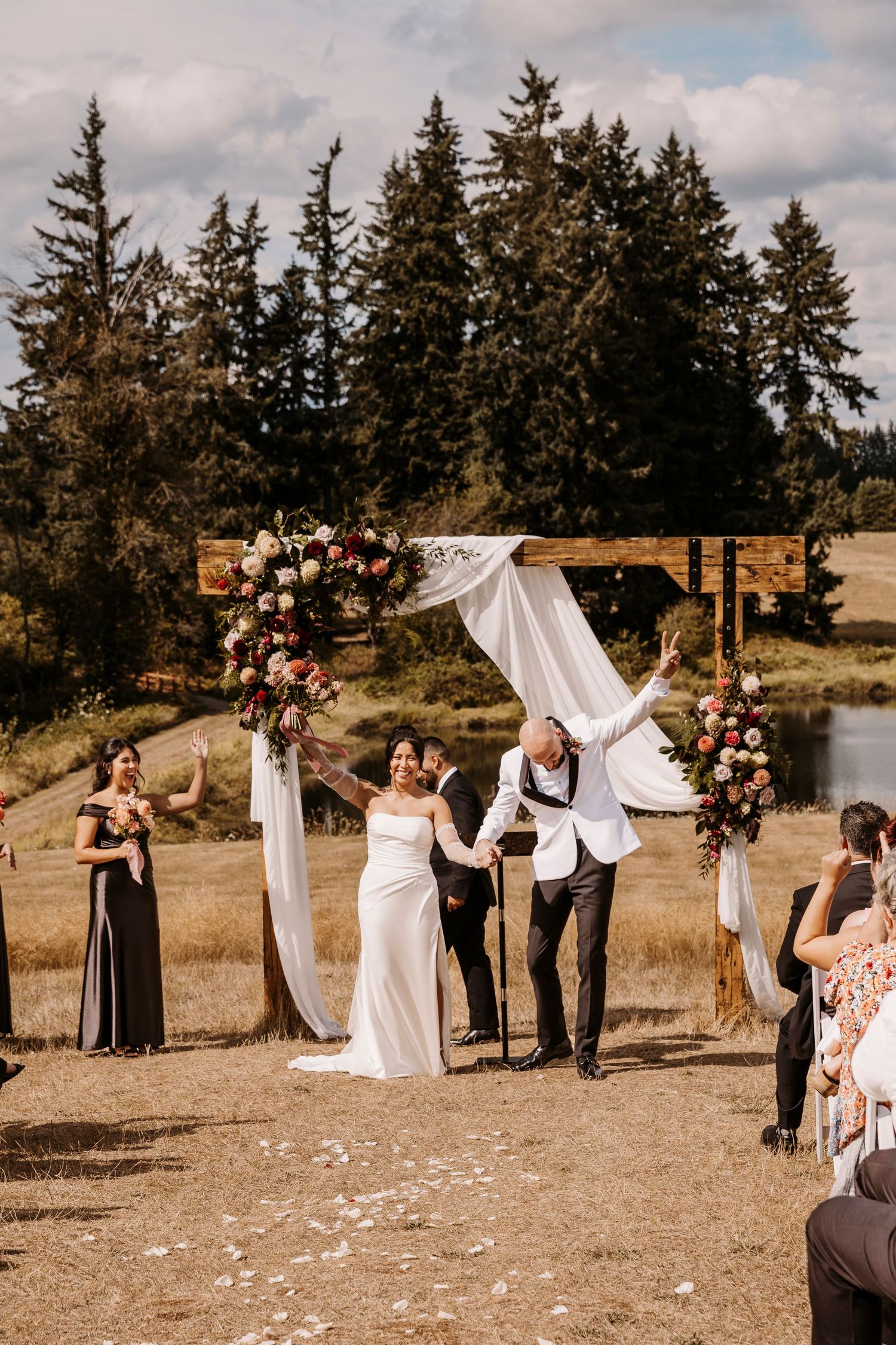 Outdoor wedding ceremony setup with white draped arch and floral arrangements against a backdrop of evergreen trees.