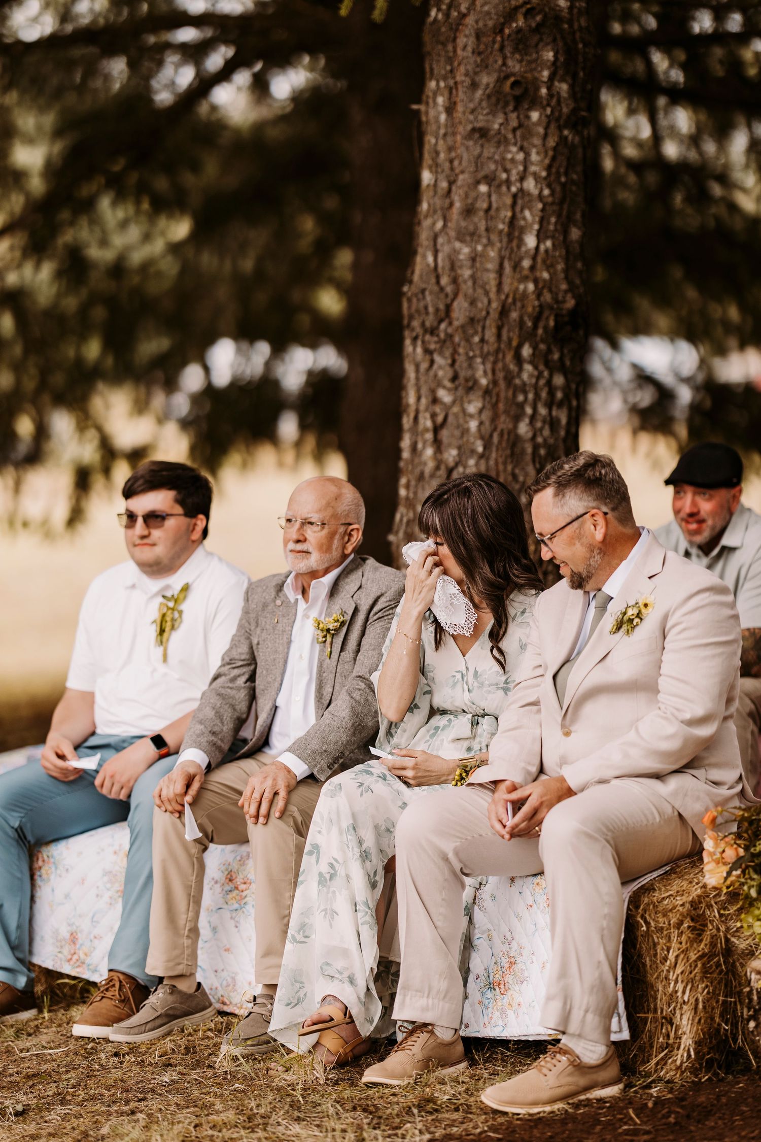 Wedding guests seated on hay bales under pine trees wearing light colored formal attire.