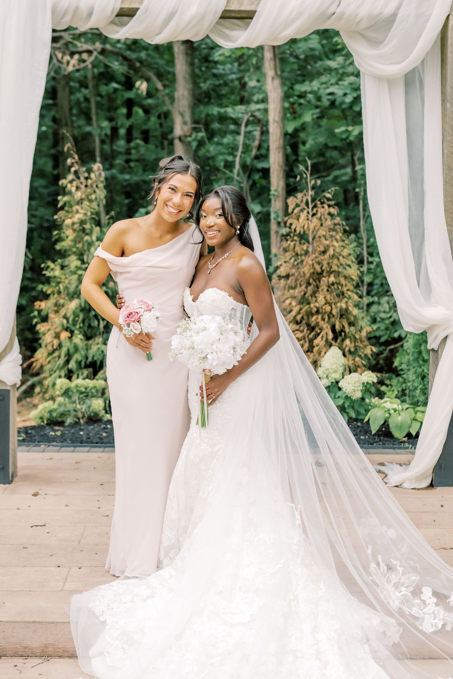 Bride holding white bouquet and bridesmaid in off-shoulder blush gown smile under white draped wedding arch.