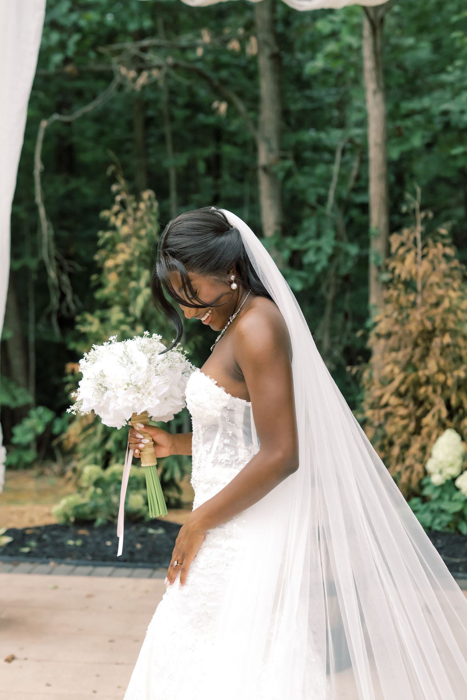 Black bride in strapless lace wedding gown holds white floral bouquet with flowing veil in garden setting.