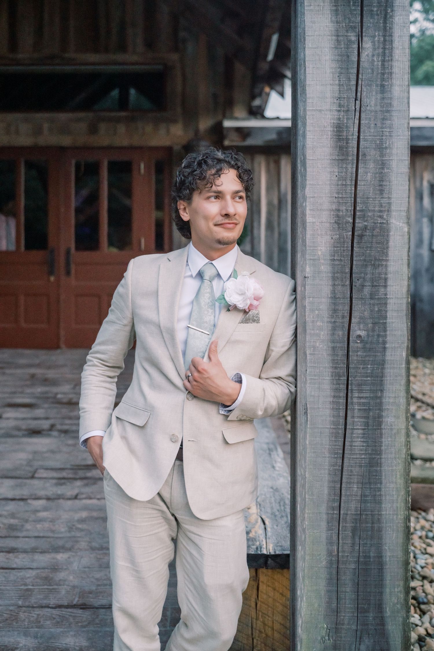 Groom in tan suit with floral boutonniere and blue tie leans against a wooden post at a rustic barn venue.