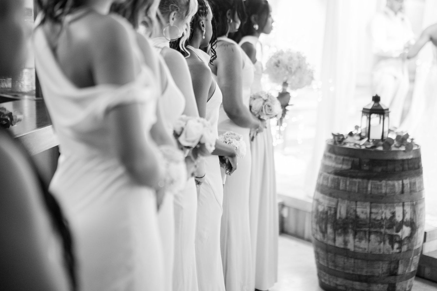 Bridesmaids in white gowns holding bouquets stand in a row near a rustic wooden barrel at a wedding.