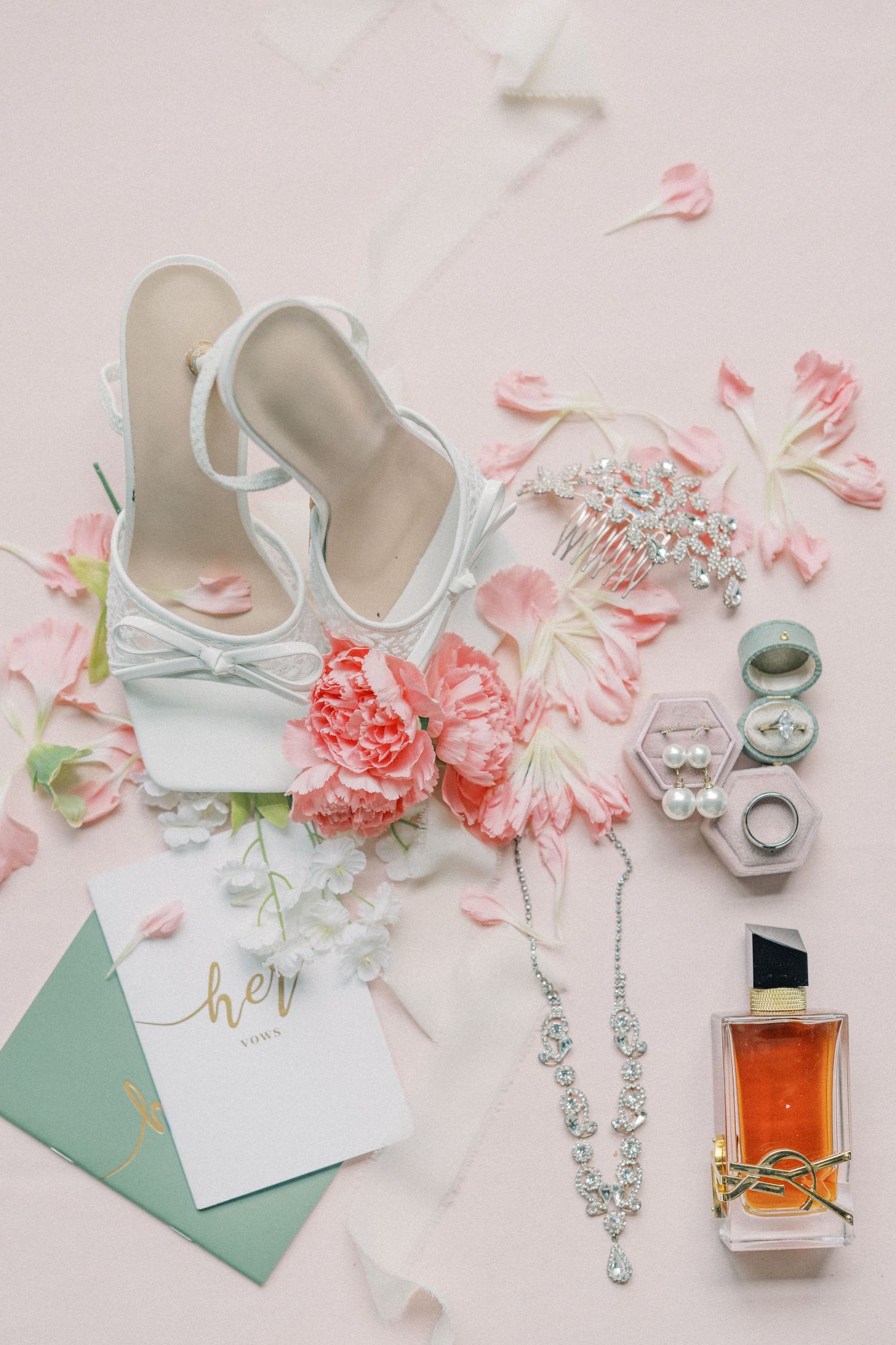Bridal flat lay with white heels, diamond necklace, YSL perfume, ring box, hair comb, and pink flowers on blush background.