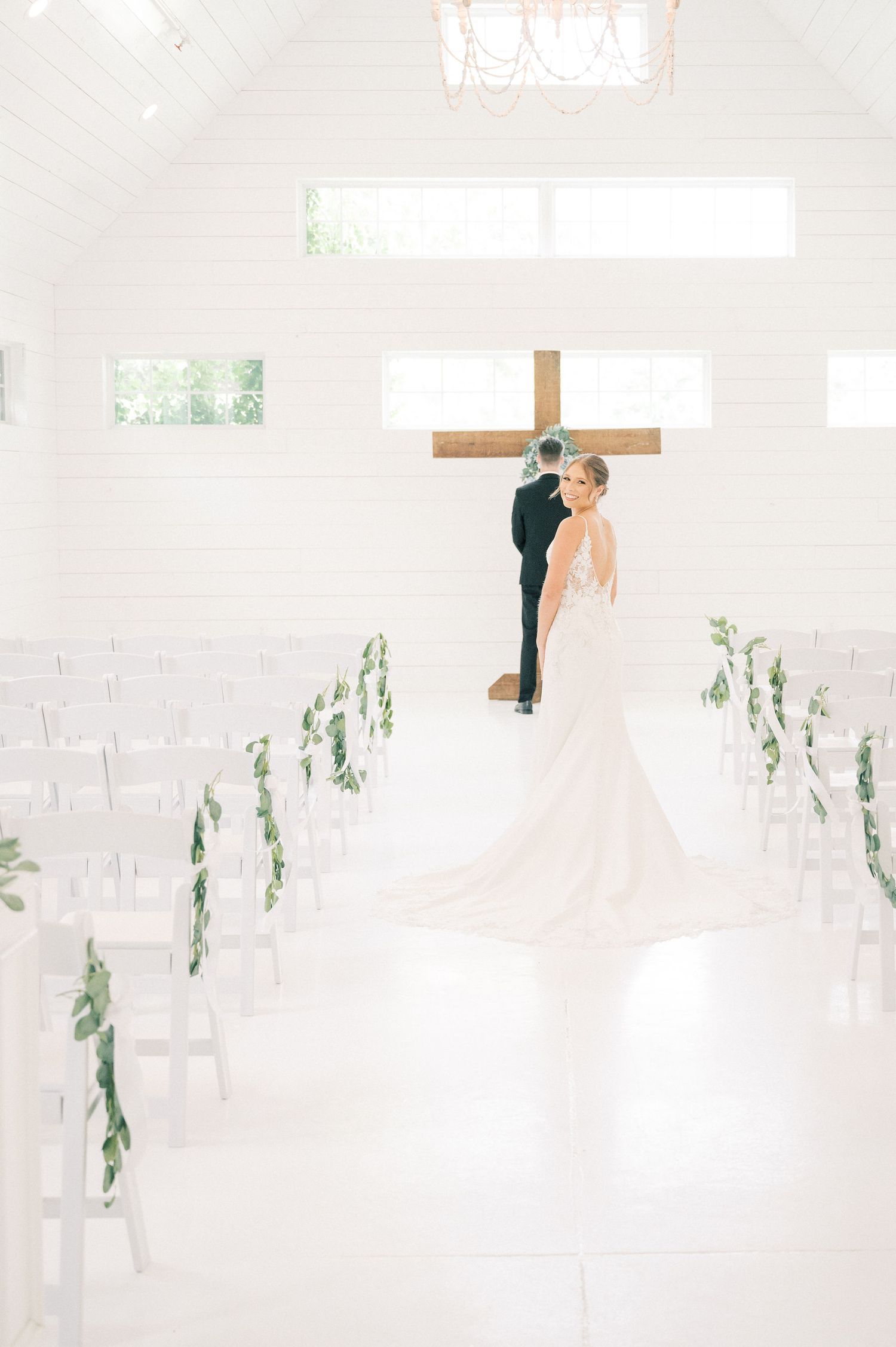 A wedding ceremony setup in a bright white chapel with greenery decorating the aisle chairs.