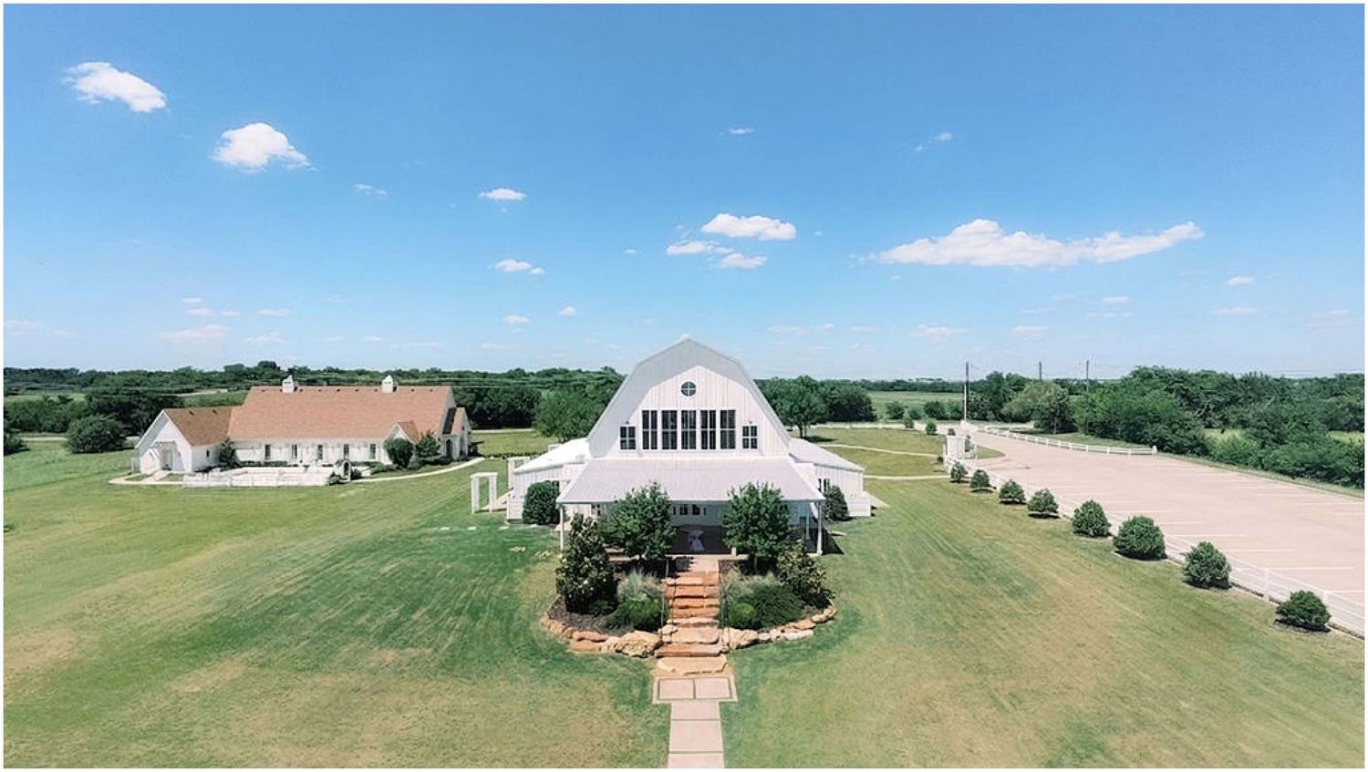 Aerial view of a white barn wedding venue surrounded by lush green lawns and manicured landscaping on a sunny day.
