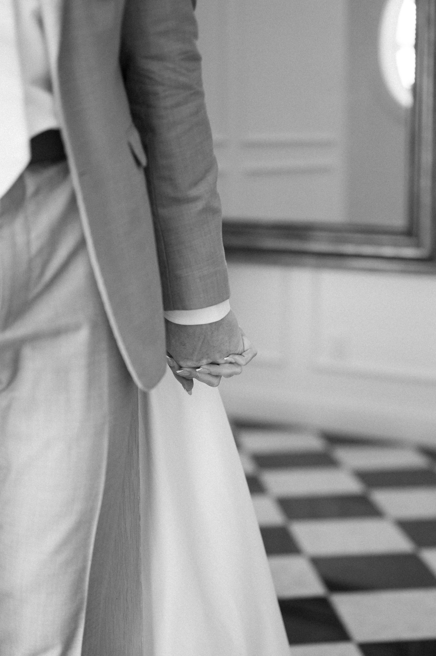 Black and white photo of a bride and groom holding hands, showcasing elegant wedding attire on a checkered floor.