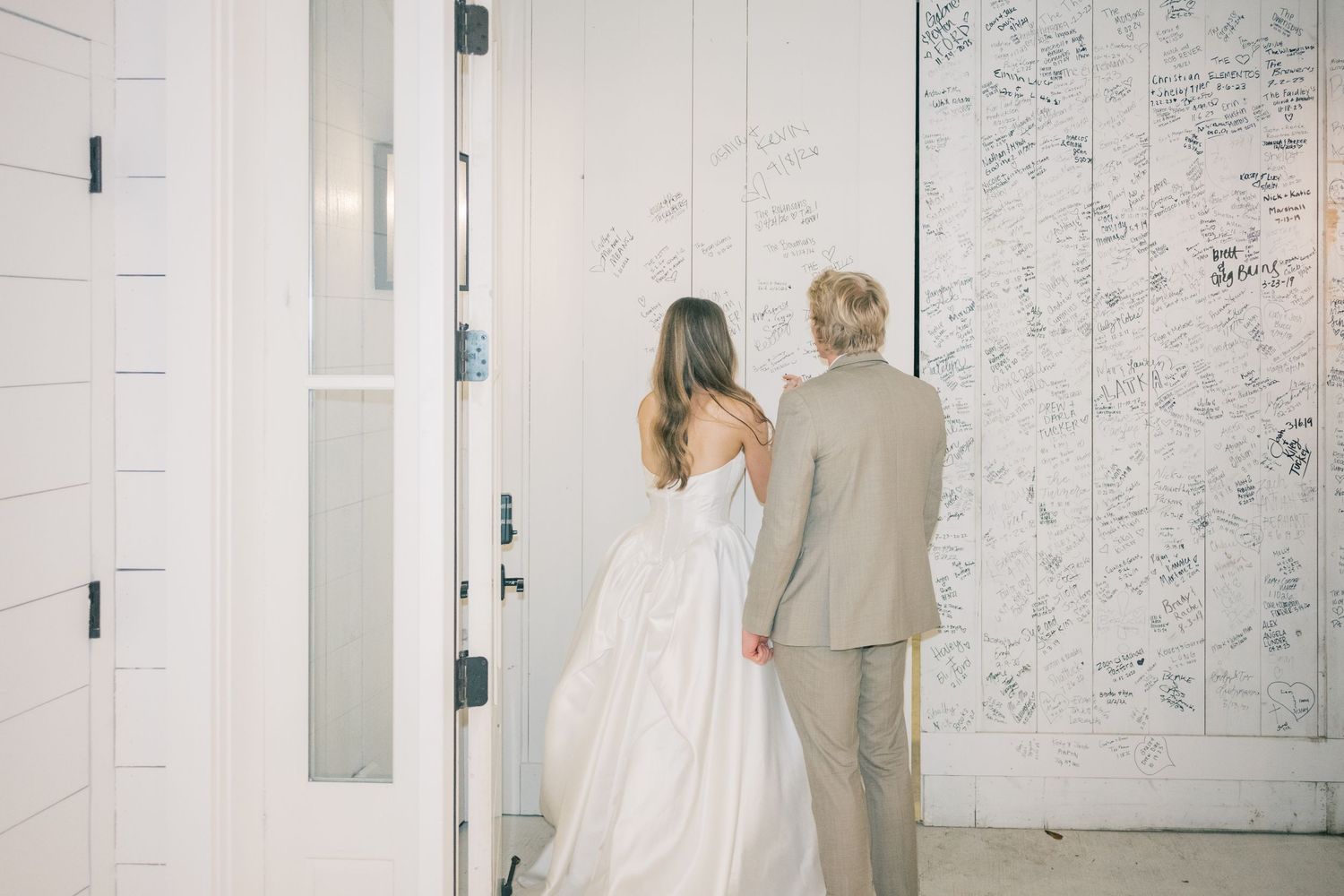 Bride and groom signing a large white wedding guest book wall, dressed in wedding attire.