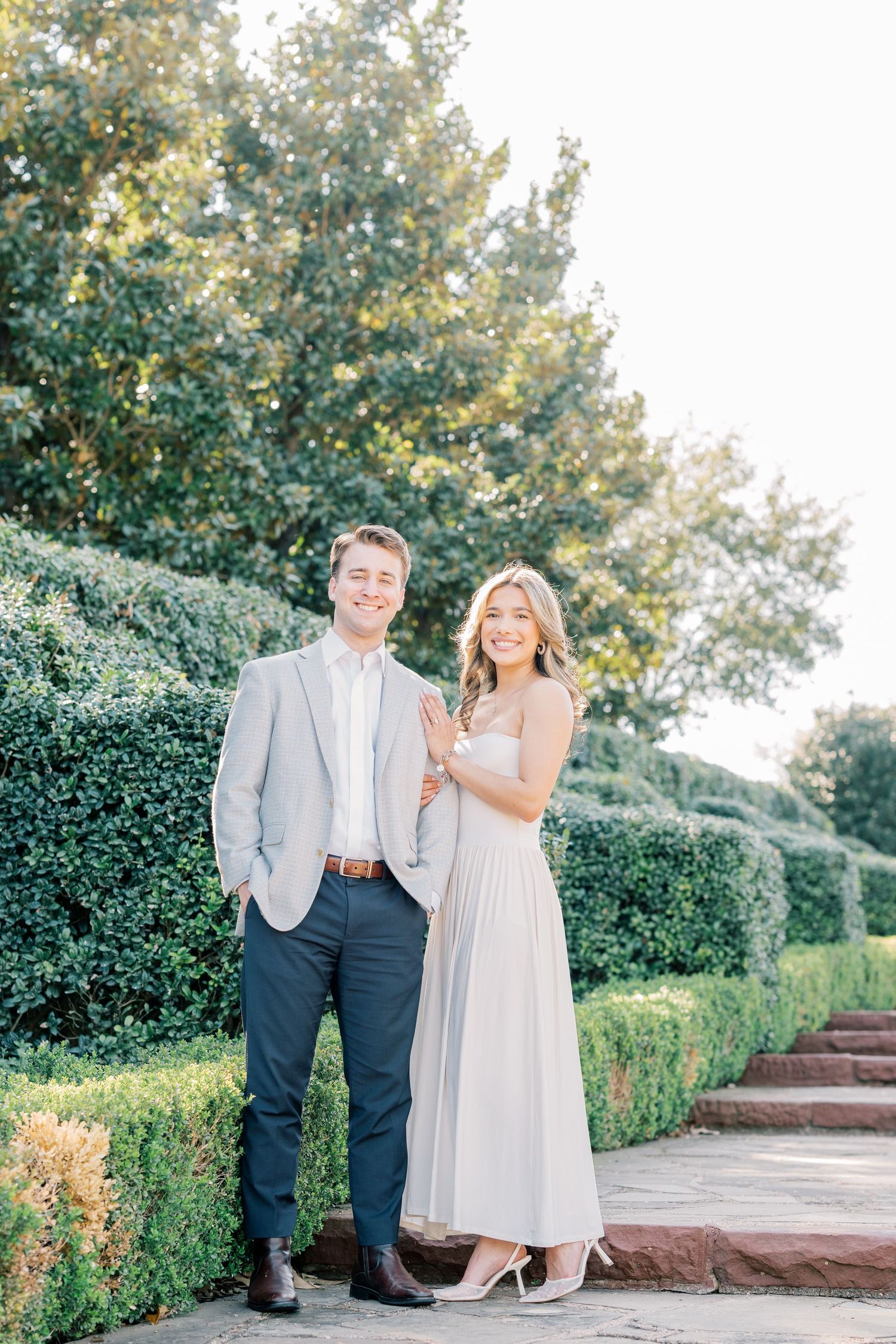 Couple in light colored formal attire pose together in a garden setting during golden hour.