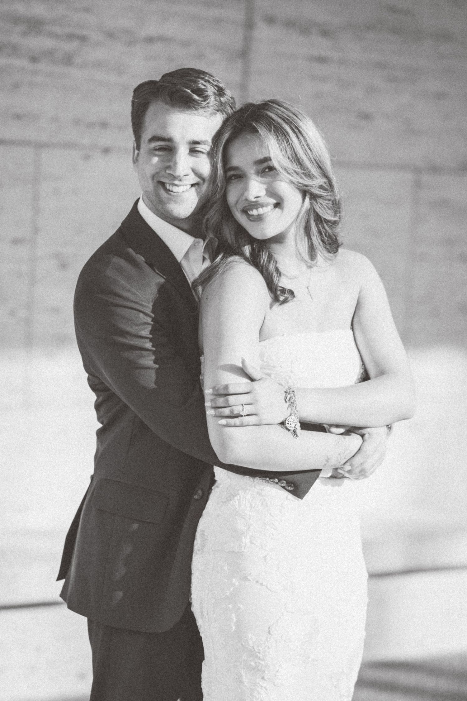 A black and white wedding portrait of a couple embracing, with the bride in a fitted lace gown and groom in dark suit.