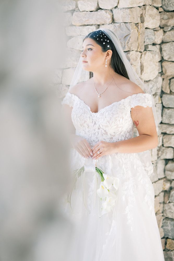 Soft portrait of bride in lace dress with pearl headpiece against rustic stone backdrop.