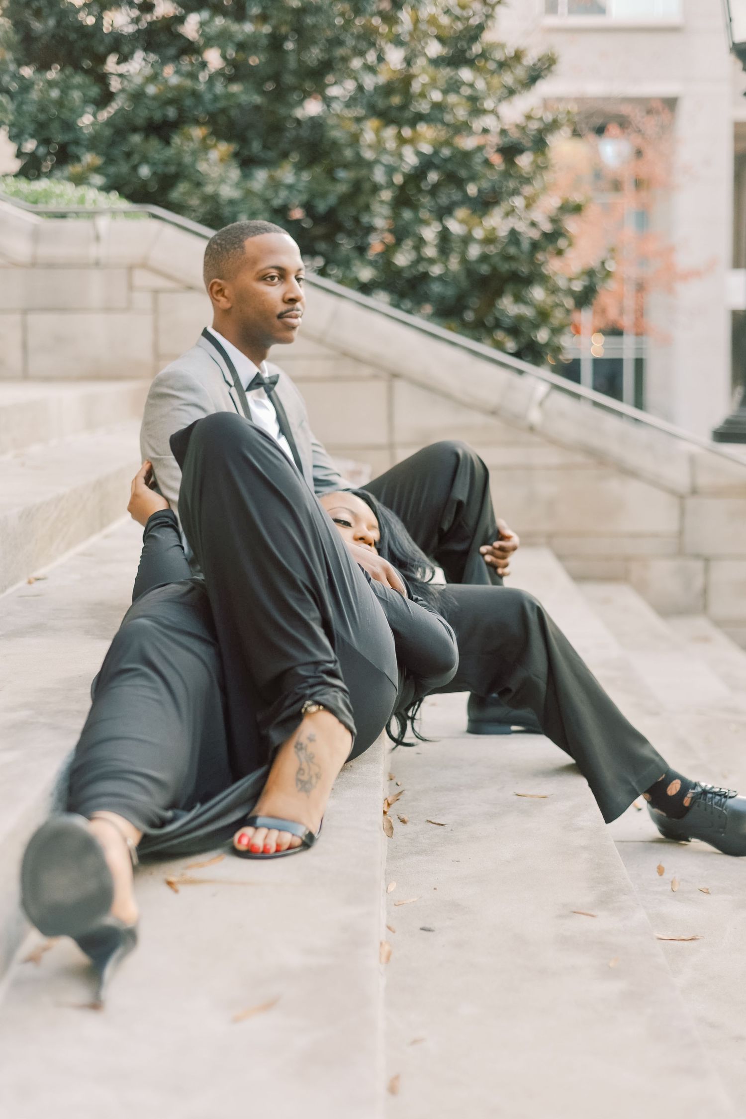 Elegant couple in formal attire sitting on stone steps outdoors, man in gray suit with bow tie, woman in black dress.