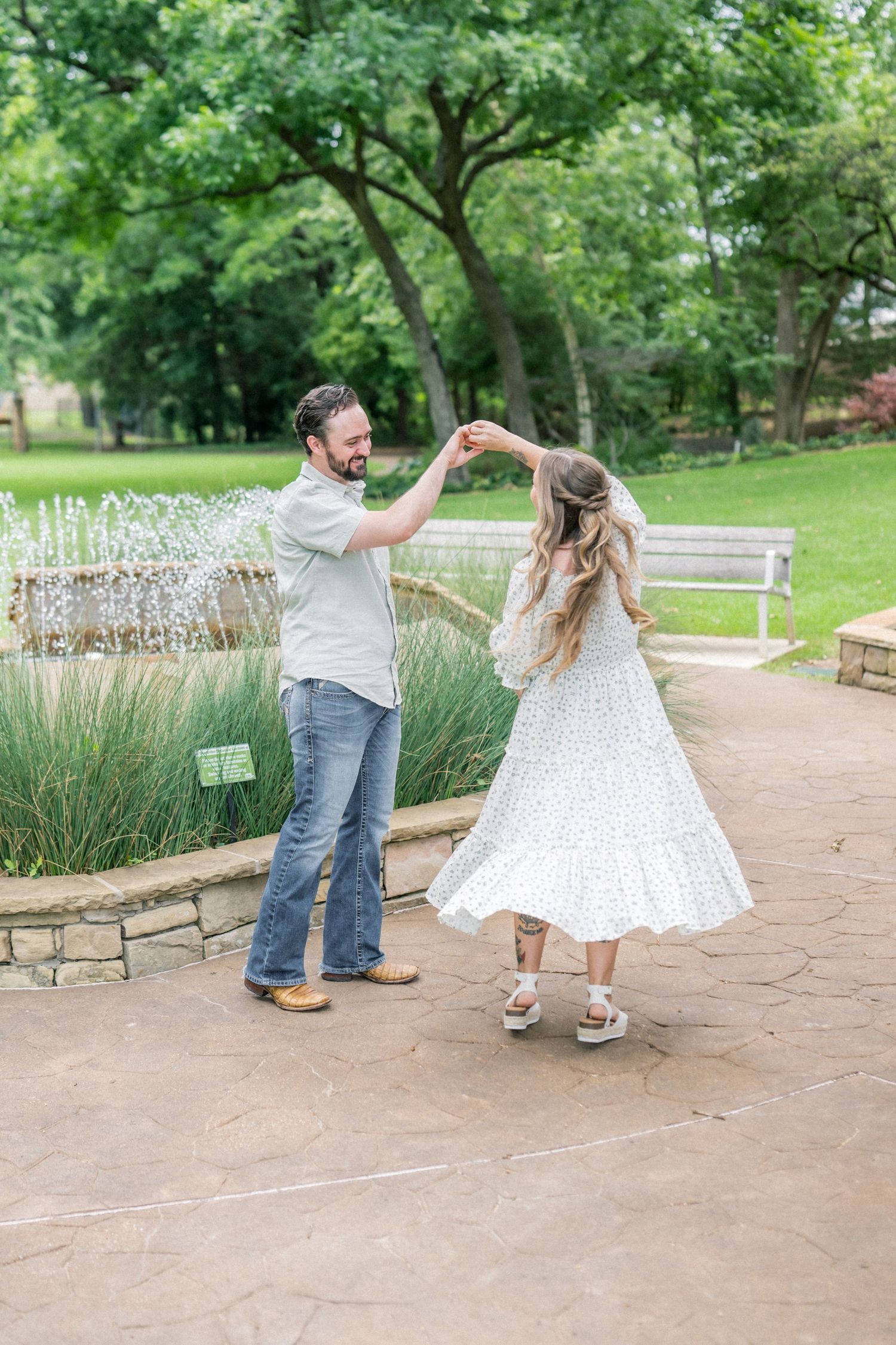 A couple dances joyfully near a fountain in a lush green park, man in jeans spinning woman in white floral dress.