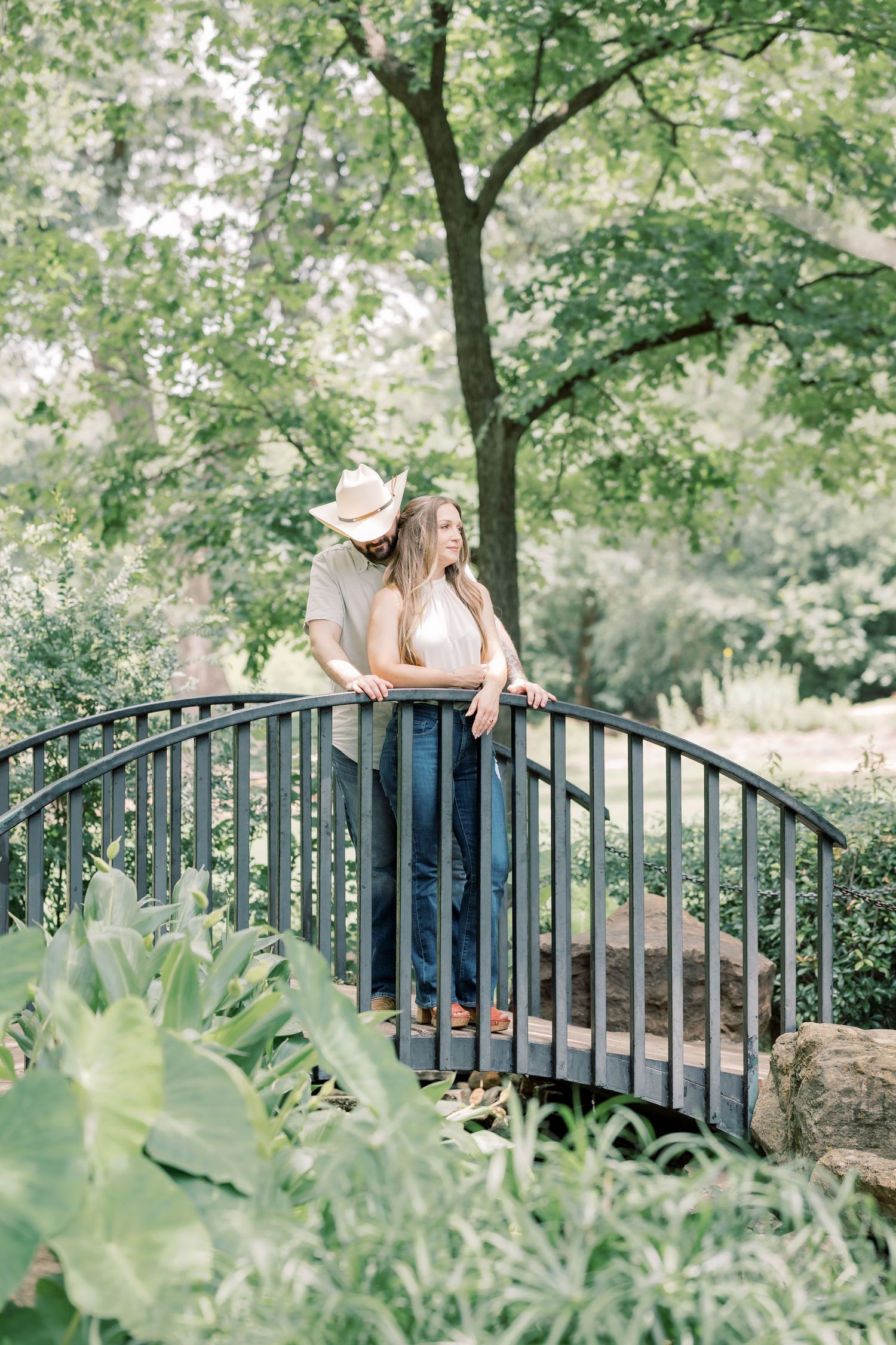 Couple standing on a curved garden bridge surrounded by lush green trees, man in cowboy hat embracing woman in jeans.