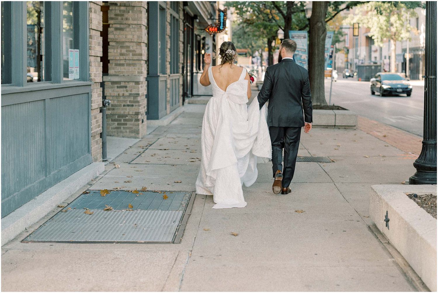 Couple in wedding attire strolling down an urban street with storefronts.
