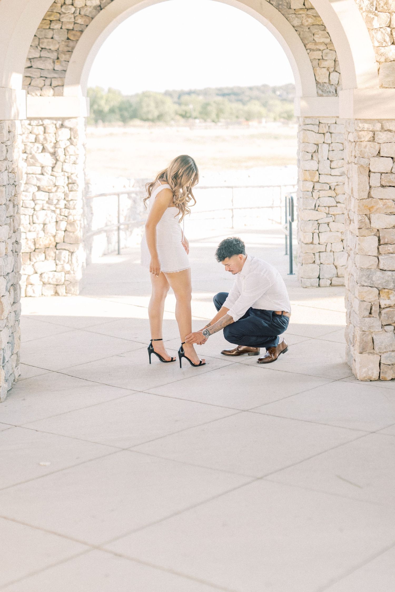 Man kneeling to adjust womans heel under stone arch during engagement photo session.