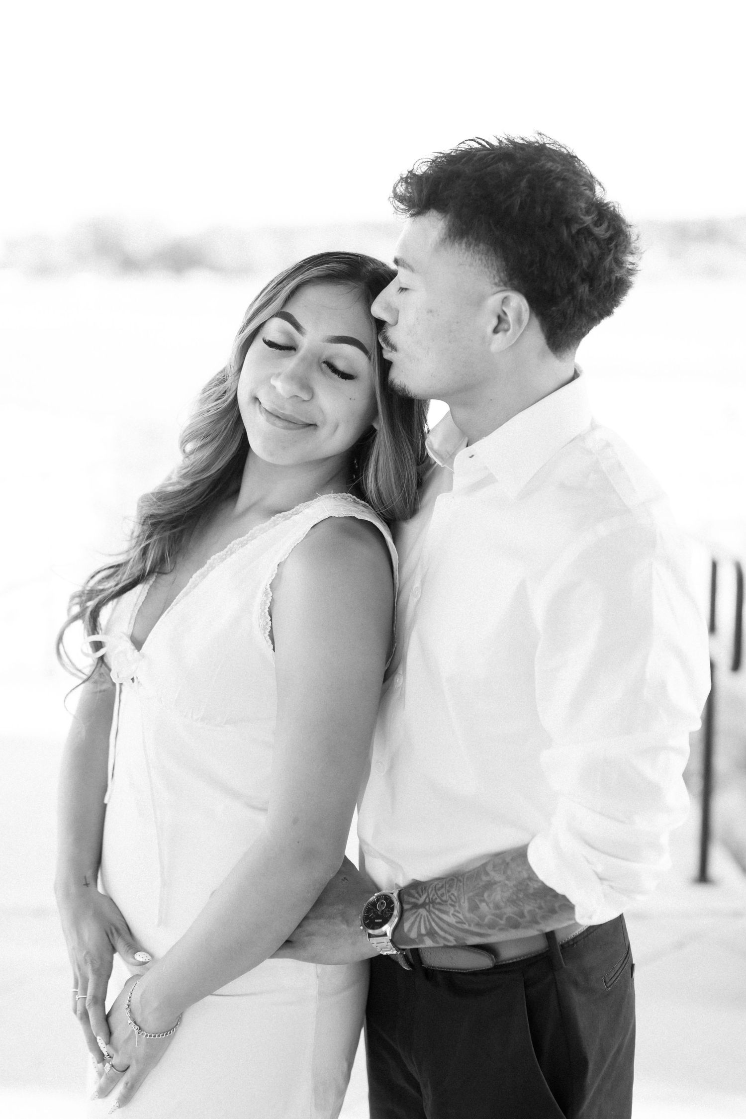 Black and white photo of a couple embracing, man kissing woman on cheek as she smiles softly.