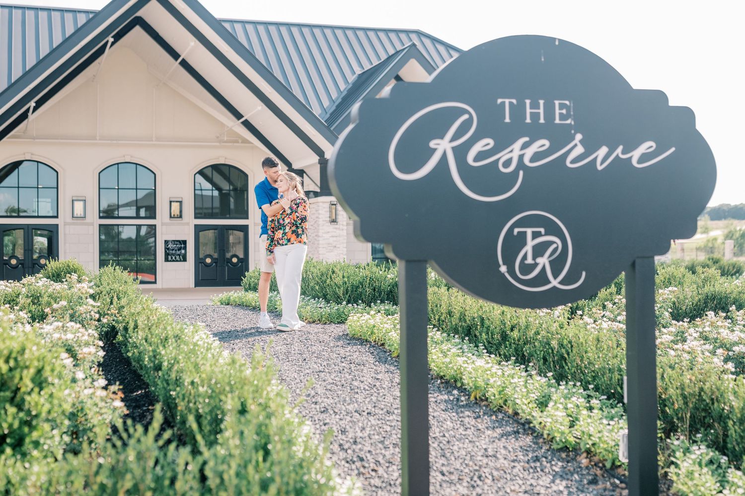 A couple kisses near The Reserve venue sign surrounded by manicured hedges and white blooms.